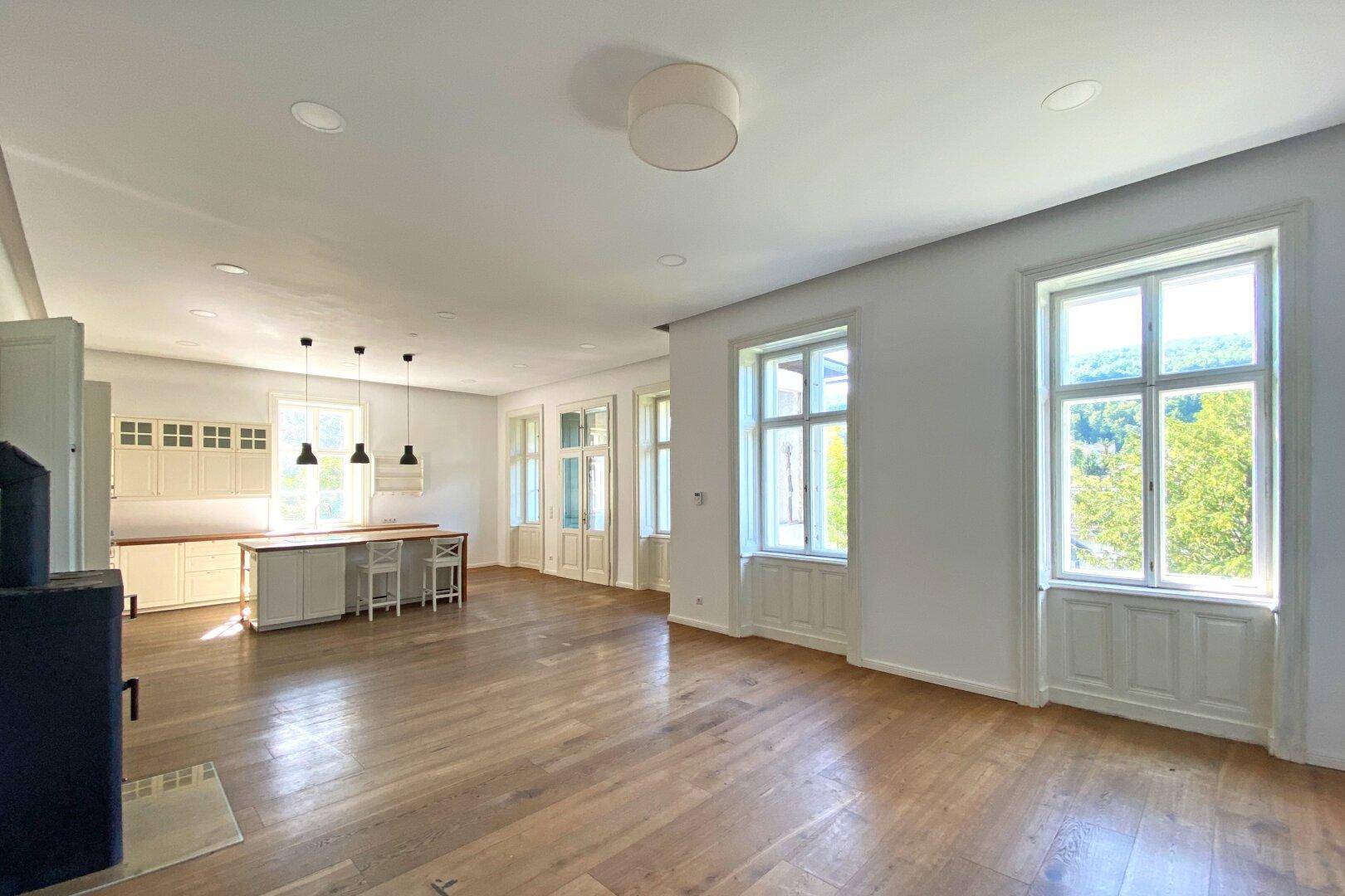 Offener Wohnbereich mit integrierter Küche, Holzboden und großen Fenstern mit Blick ins Grüne.