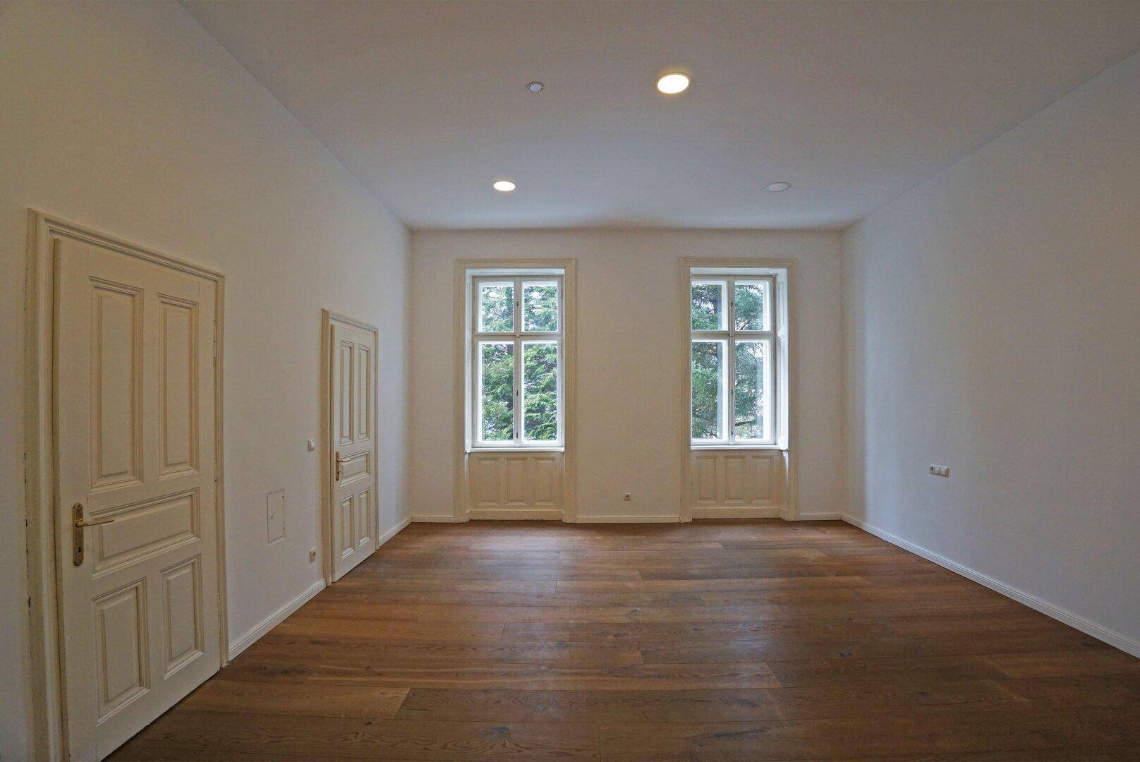 Geräumiges Zimmer mit Holzboden und hohen Fenstern, die viel Licht hereinlassen.