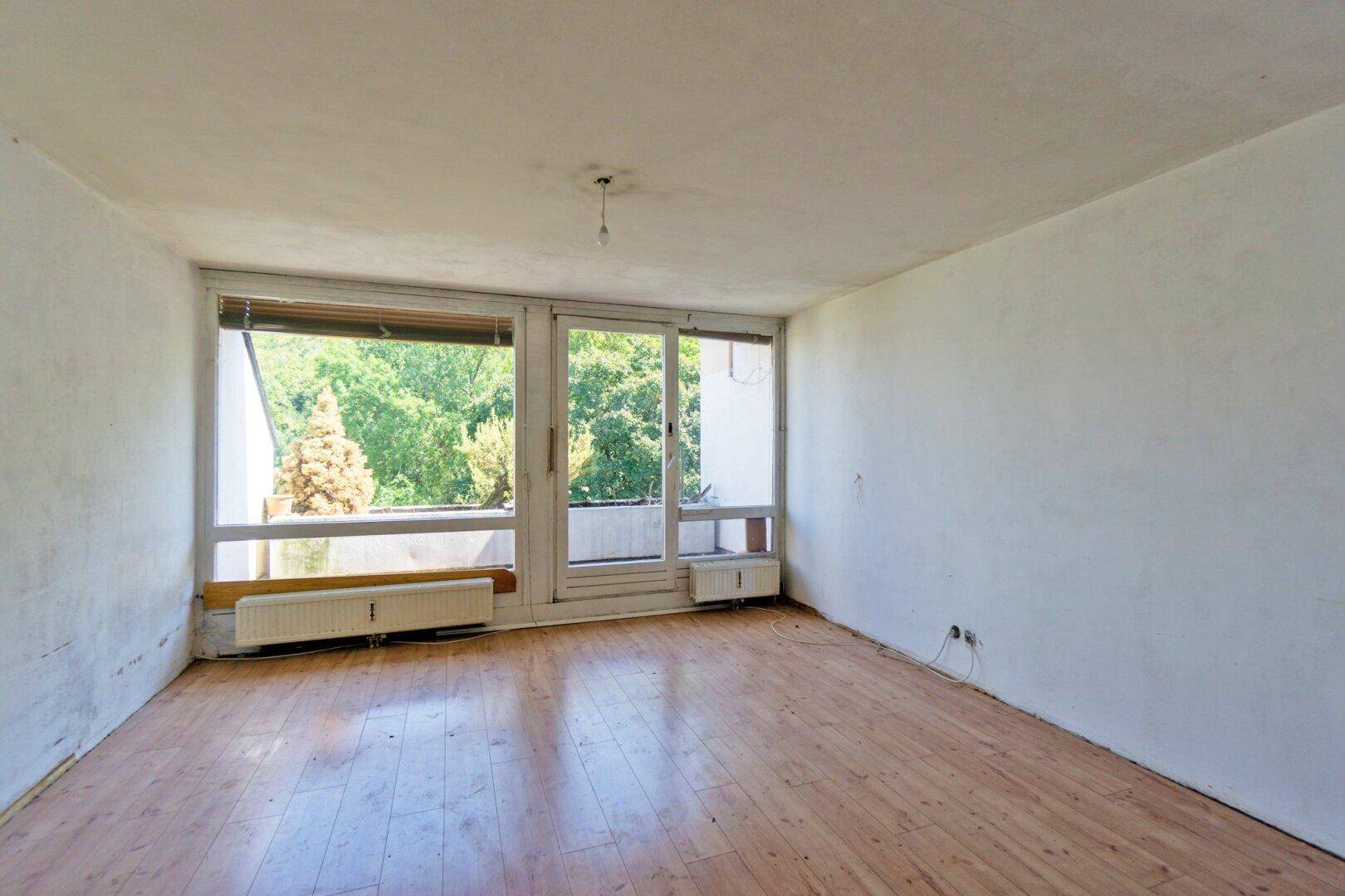 Helles, leeres Zimmer mit großen Fenstern und Blick ins Grüne, bereit zur Renovierung.