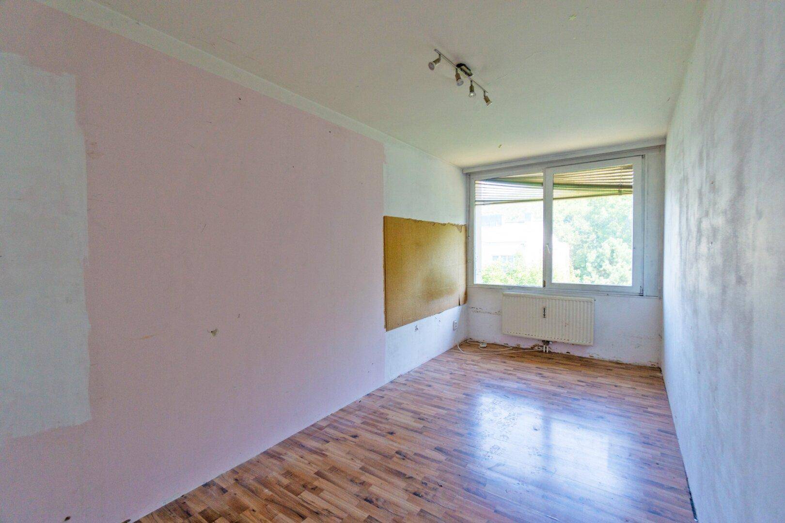 Leeres Zimmer mit Laminatboden und Fenster, das auf eine umfassende Renovierung wartet.