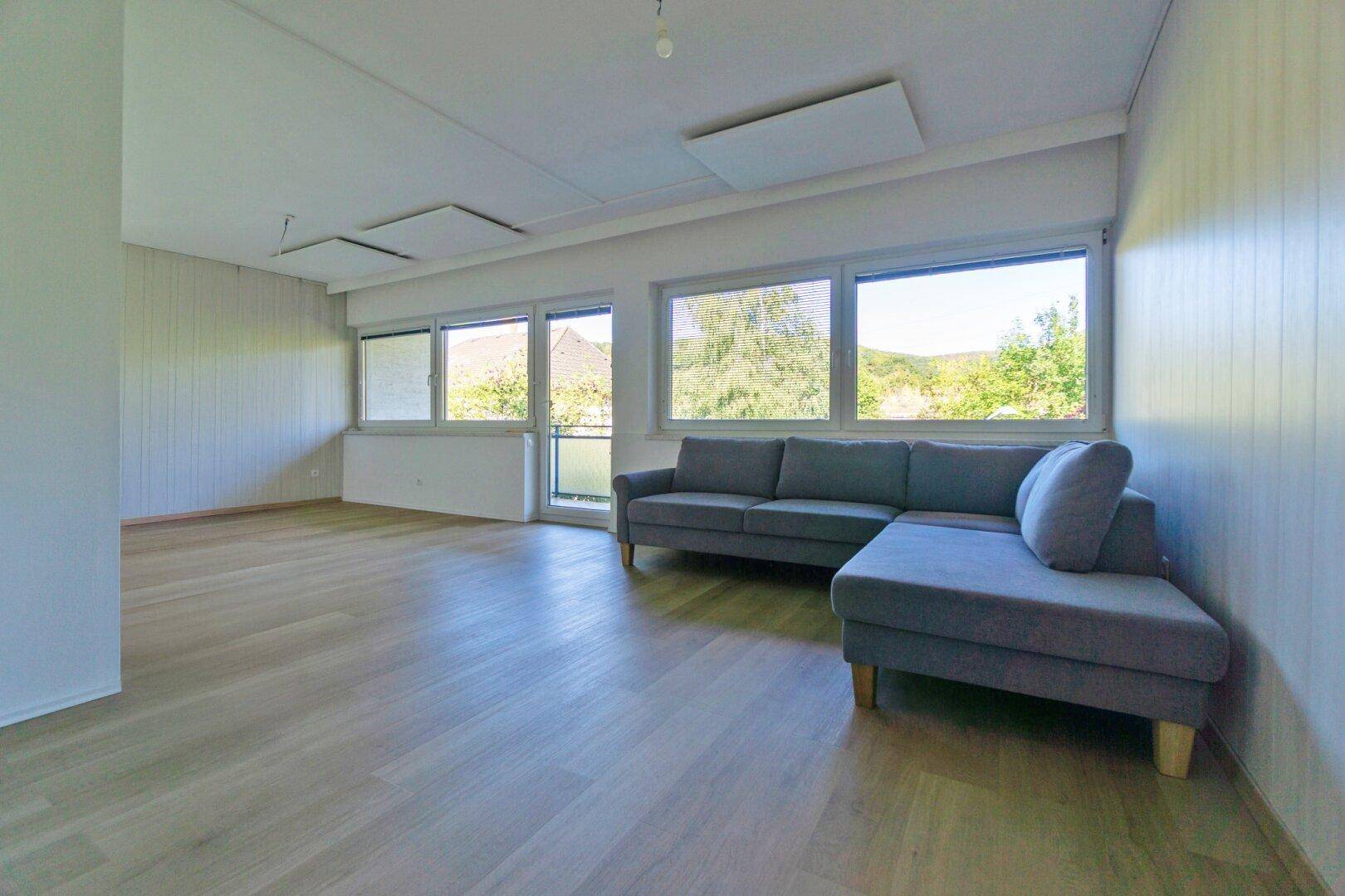 Helles Wohnzimmer mit Laminatboden und großen Fenstern, die viel Tageslicht hereinlassen.