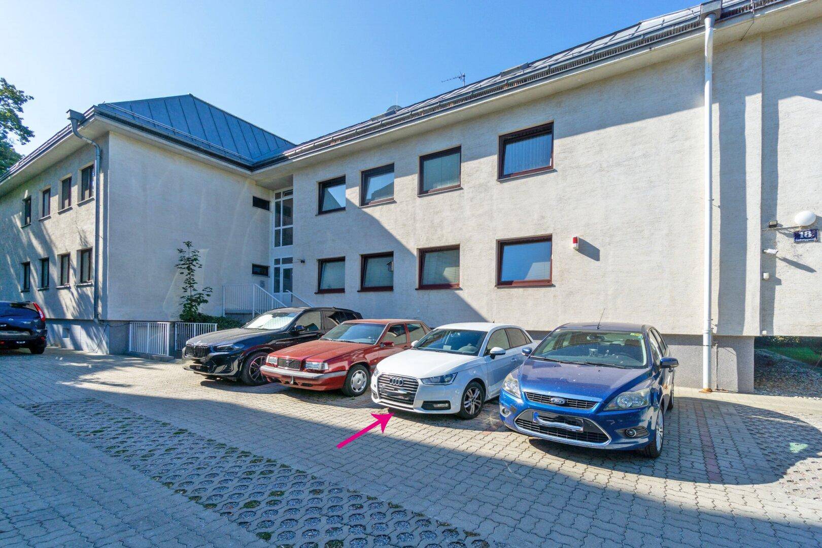 Gebäudefassade mit mehreren geparkten Autos auf dem Vorplatz an einem sonnigen Tag.