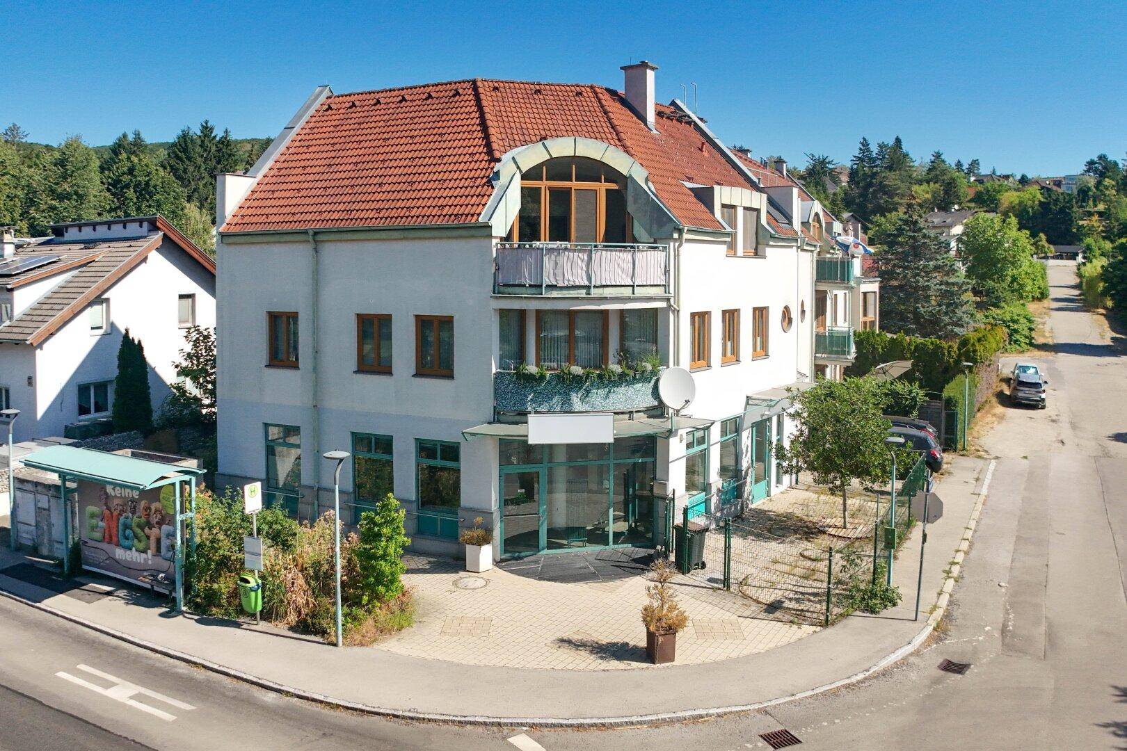Mehrstöckiges Gebäude mit rotem Ziegeldach, großen Fenstern und einem kleinen Balkon an einer Straßenecke.