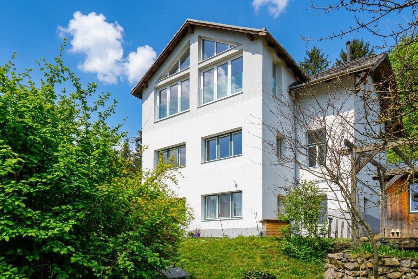 Weißes Haus mit vielen Fenstern und Giebeldach, umgeben von üppigem Grün.