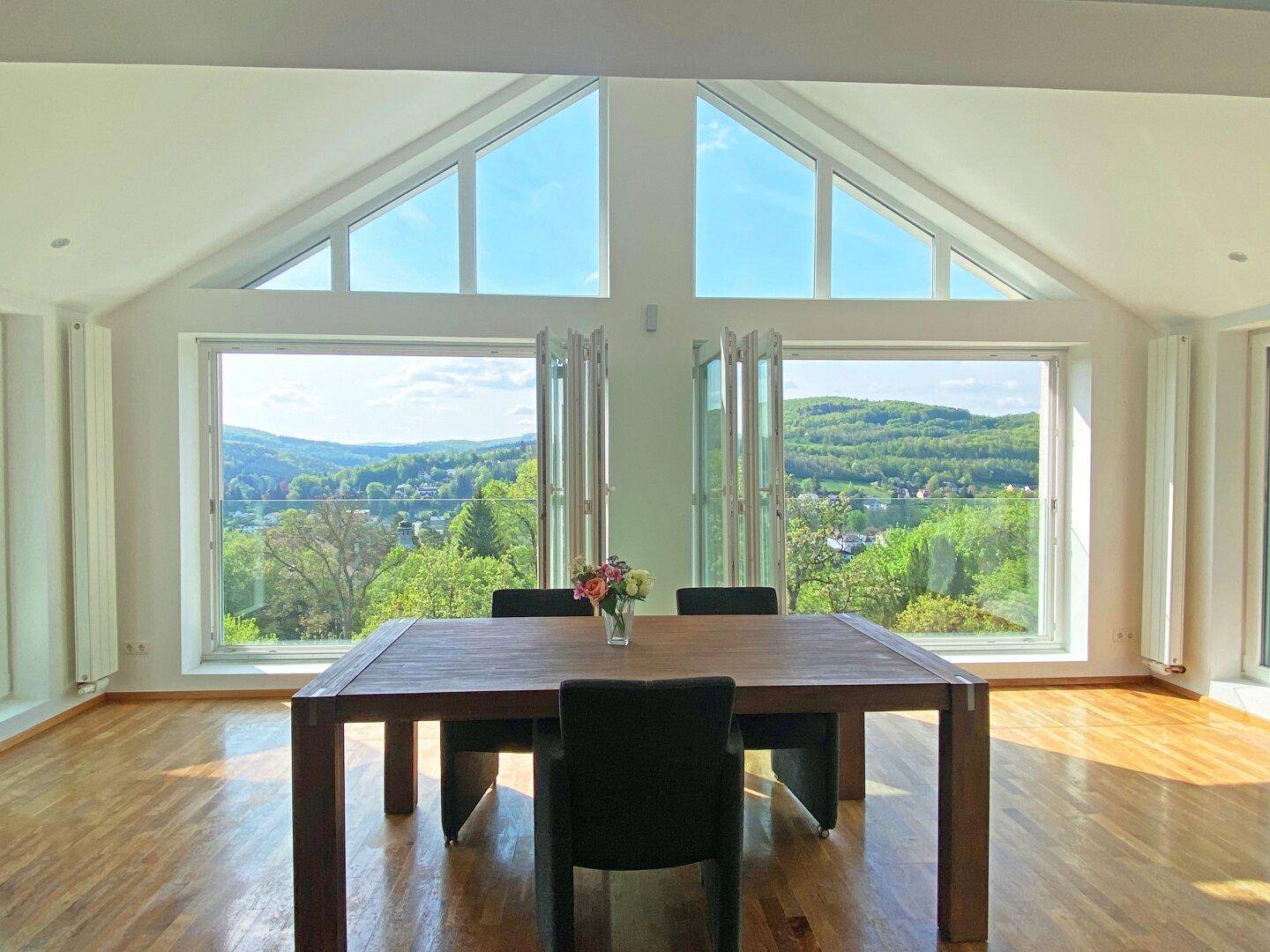 Heller Essbereich mit Holztisch und Stühlen, umgeben von großen Fenstern mit Weitblick.