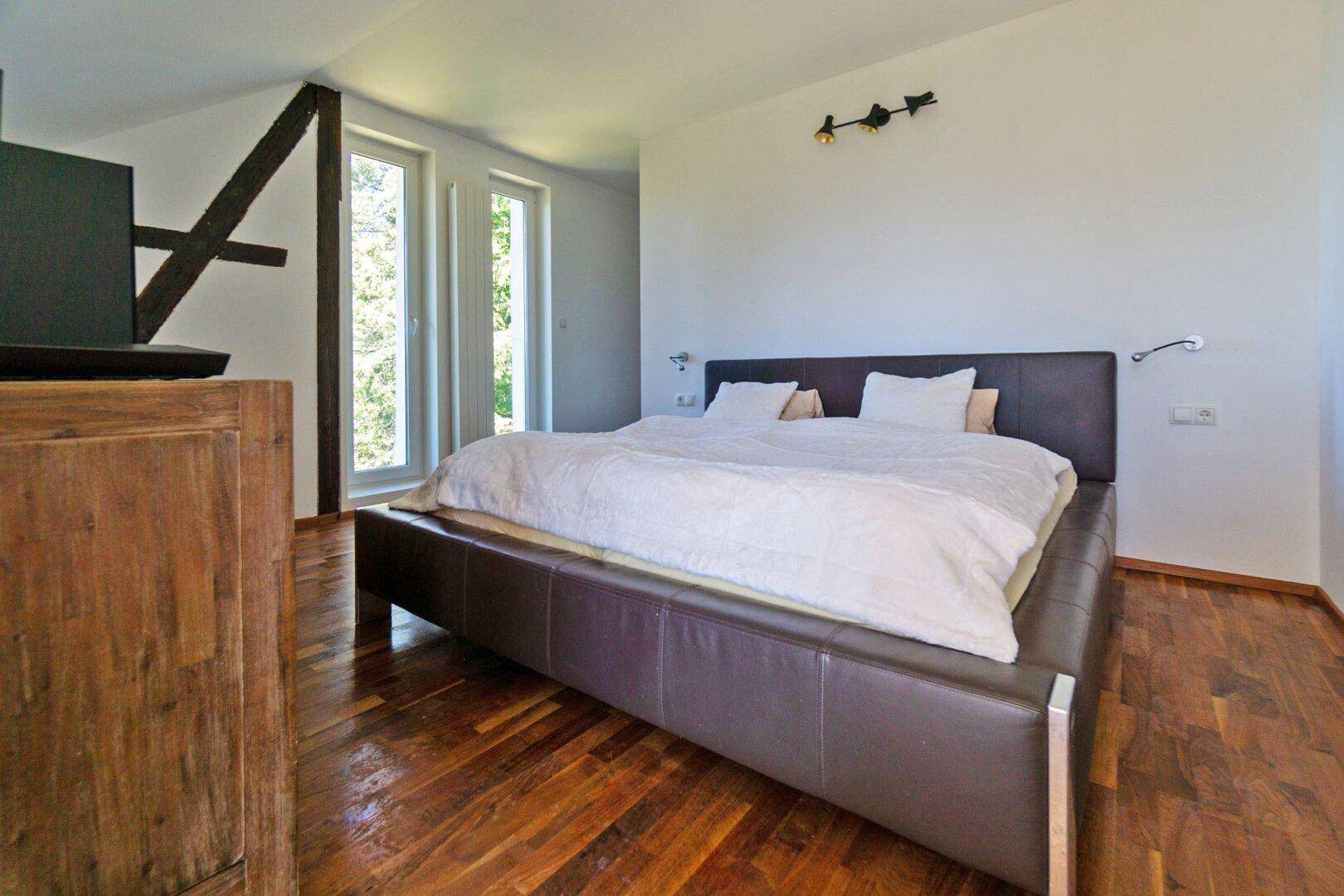 Gemütliches Schlafzimmer mit großem Lederbett, Holzboden und hohen Fenstern unter Dachschräge.