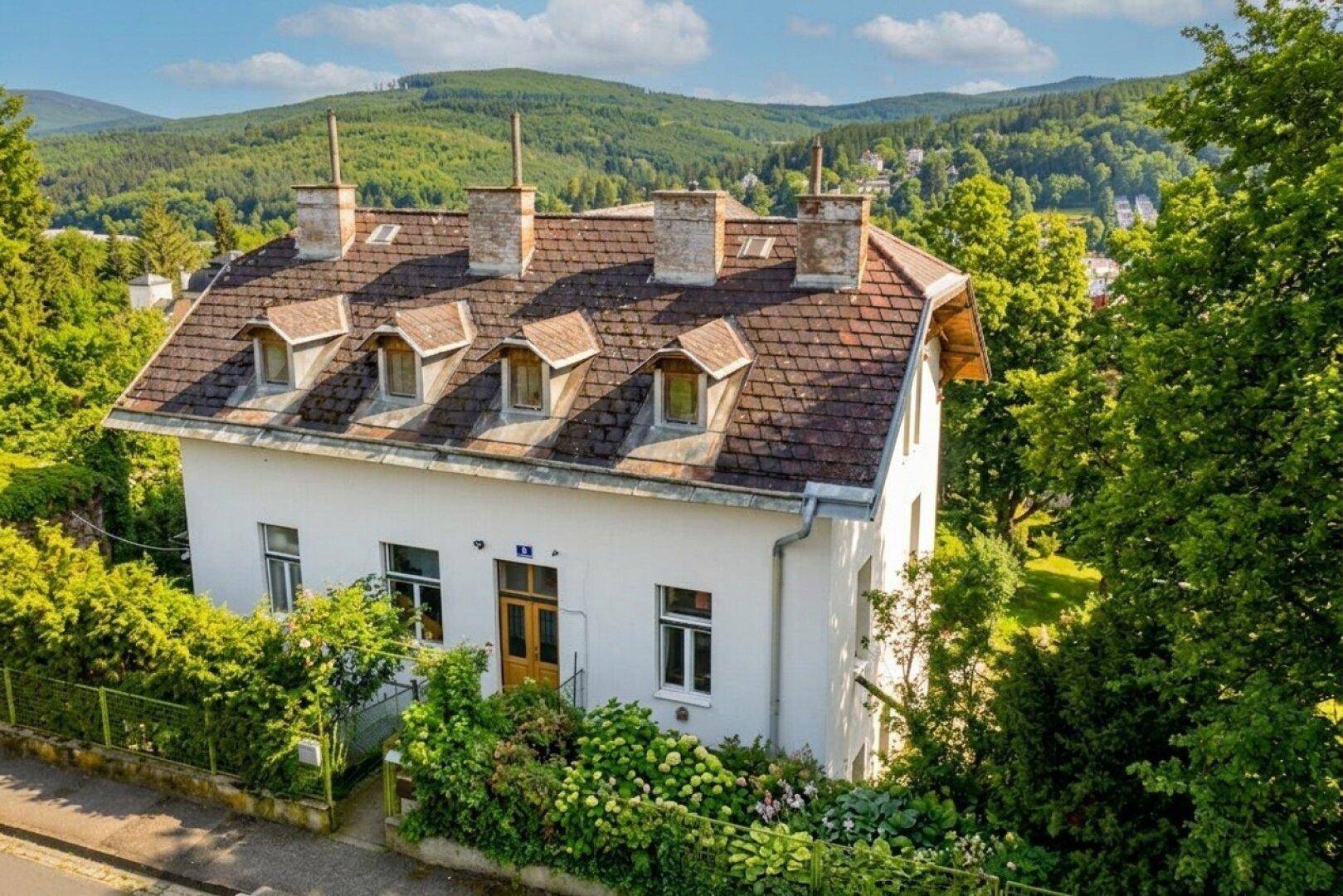 Traditionelles weißes Haus mit Ziegeldach, Gauben und üppiger Bepflanzung im Vorgarten.