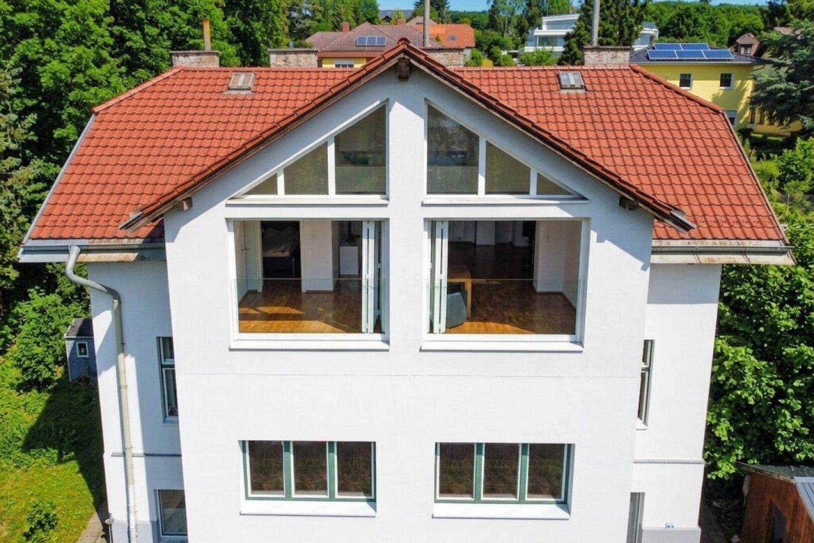 Fassade des weißen Hauses mit großen Fenstern und rotem Ziegeldach unter blauem Himmel.