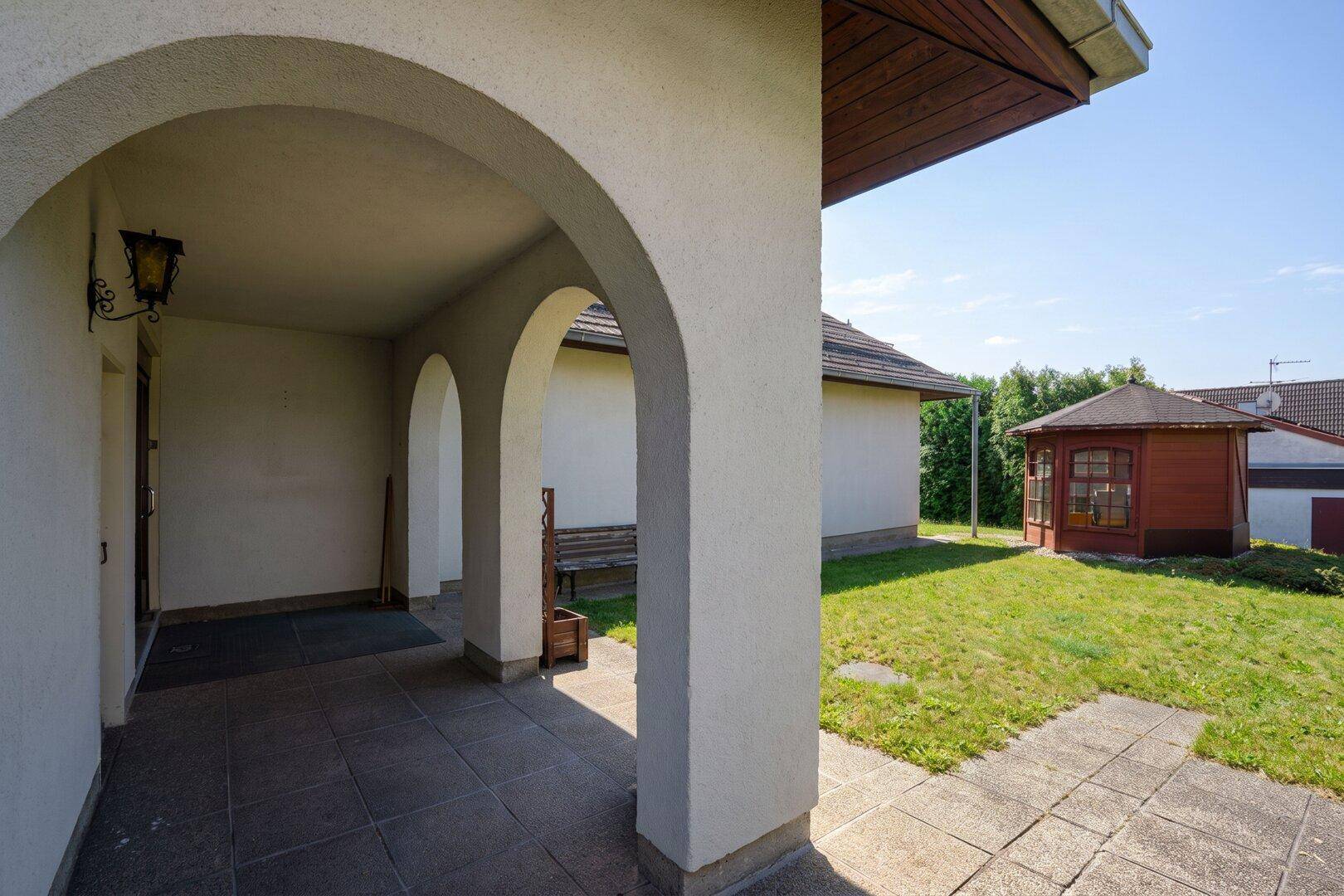 Überdachter Eingangsbereich mit Bögen und Blick auf den Garten mit Gartenhaus.