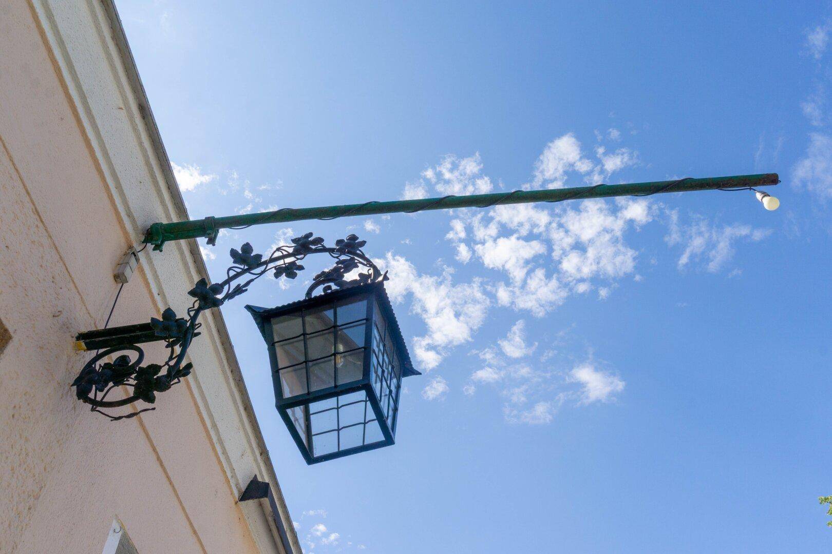 Detailaufnahme einer verzierten Außenlaterne an der Fassade eines Gebäudes vor blauem Himmel.