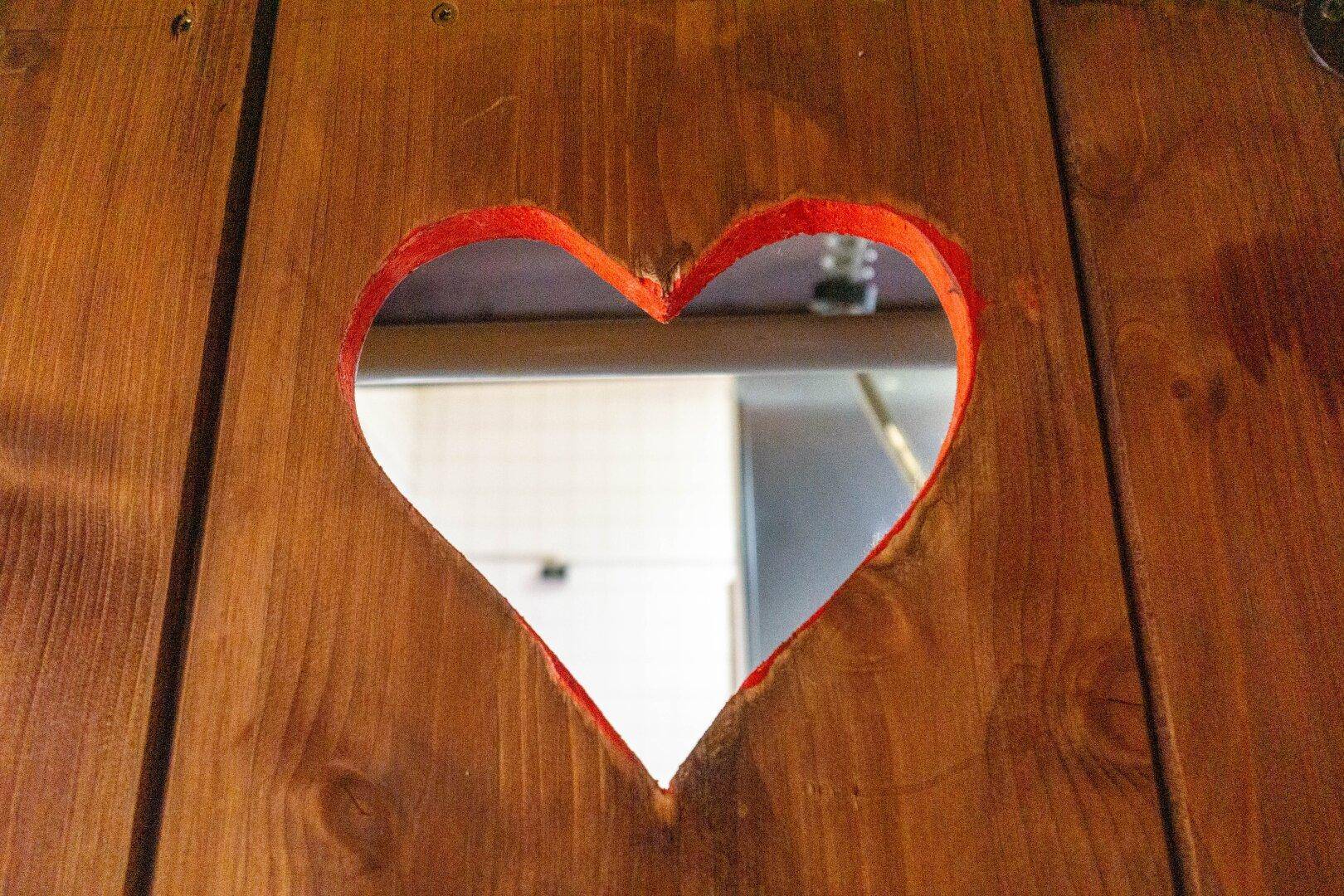 Nahaufnahme eines herzförmigen Ausschnitts in einer Holztür oder -wand, der einen Blick in einen anderen Raum freigibt.