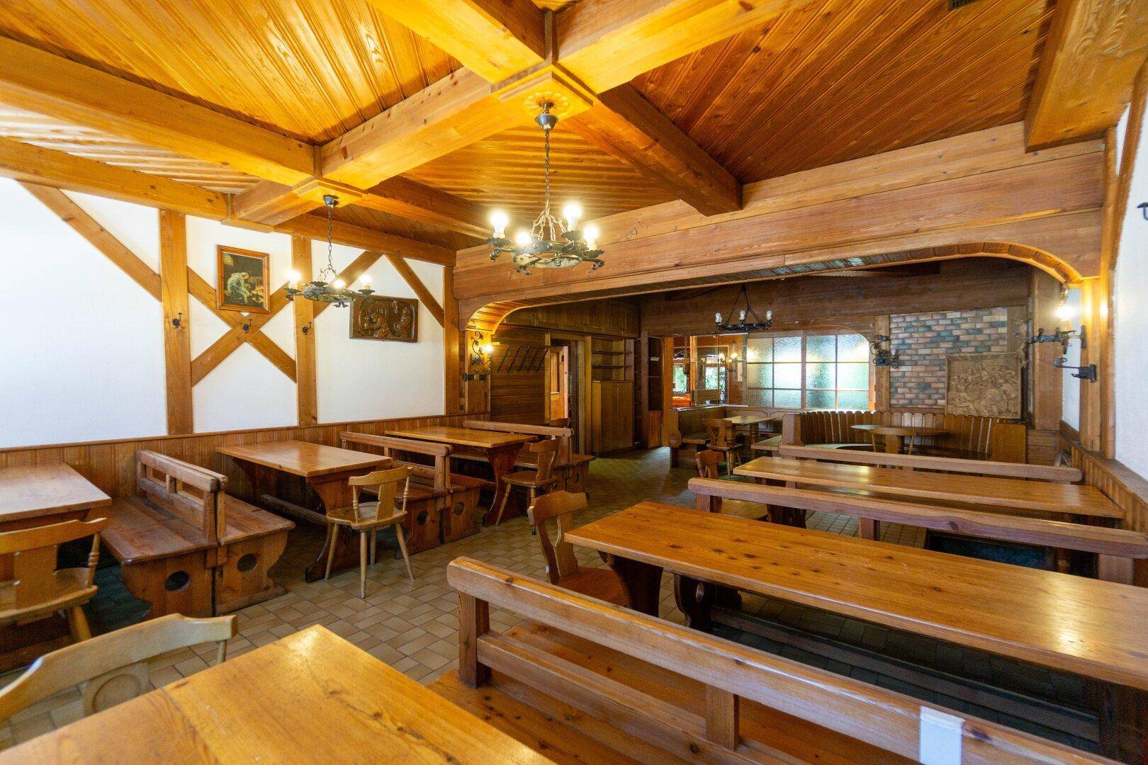 Heller Gastraum mit Holztischen und Bänken, weißen Wänden mit dekorativen Holzbalken und Holzbalkendecke.