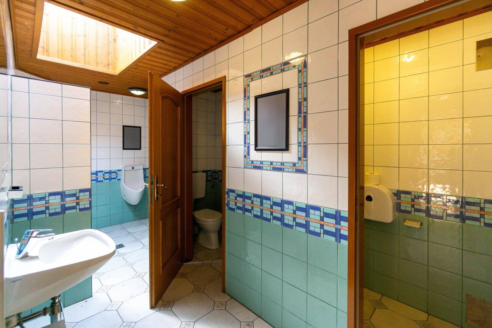 Öffentliche Toilettenanlage mit mehreren Kabinen, Urinal, Waschbecken und farbigen Fliesen an Wänden und Boden.