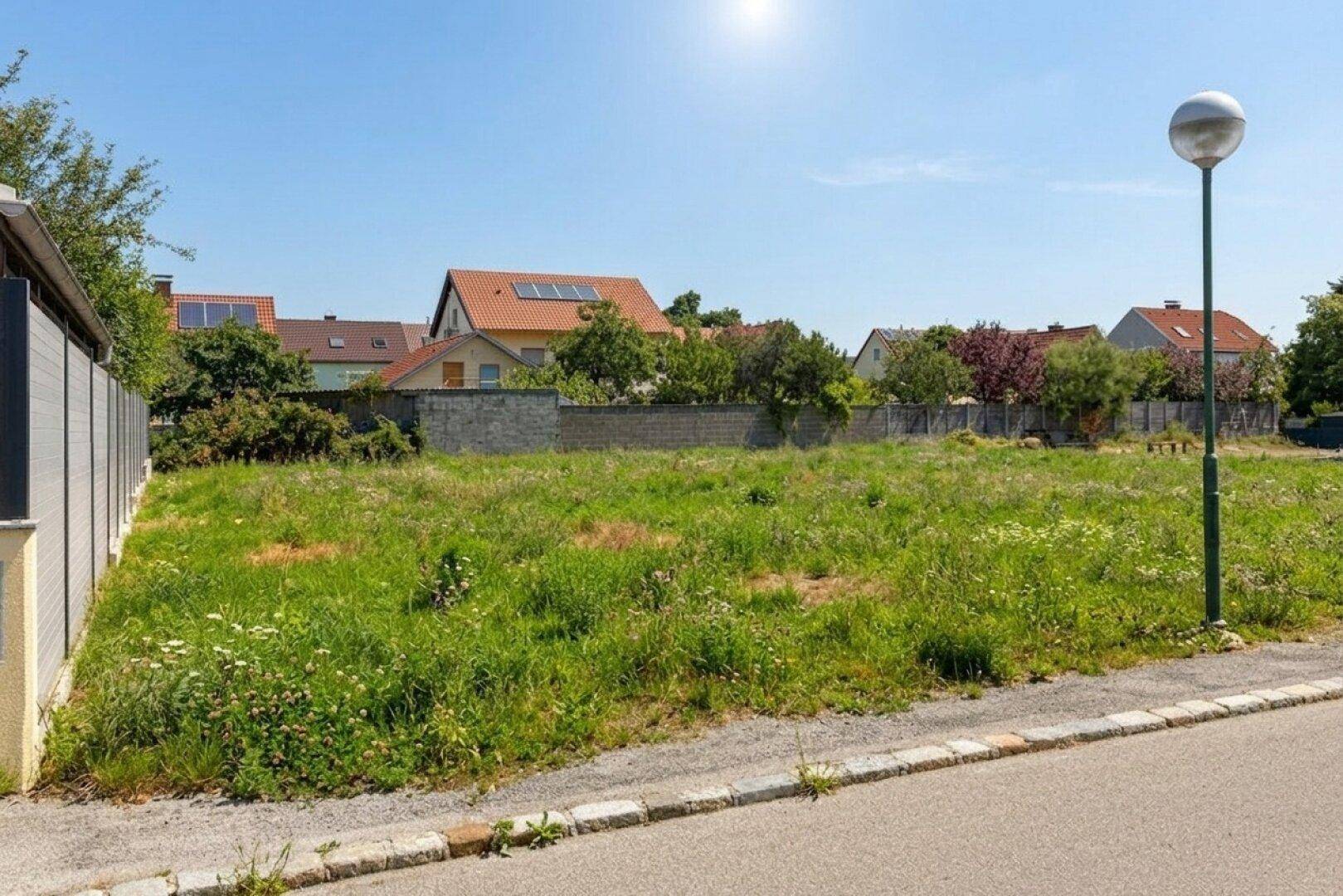Sonniges unbebautes Grundstück von der Straße aus gesehen, mit Grasfläche und angrenzenden Häusern.