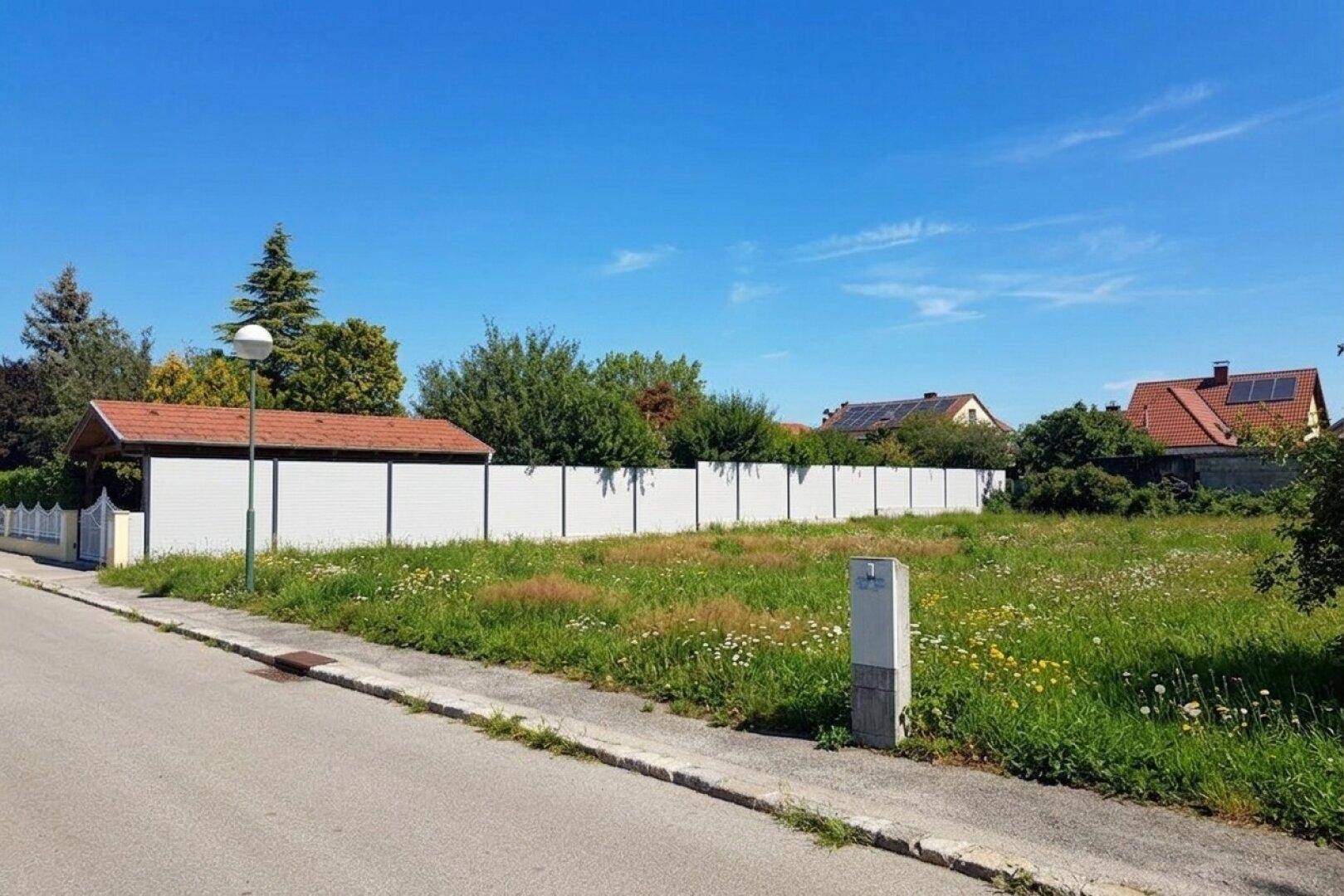 Unbebautes Grundstück mit weißem Zaun entlang der Grenze und Wohnhäusern im Hintergrund unter blauem Himmel.