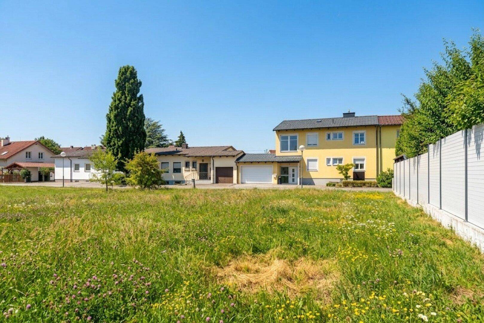 Weitläufiges unbebautes Grundstück mit Blick auf ein gelbes Wohnhaus und weitere Gebäude in der Nachbarschaft.