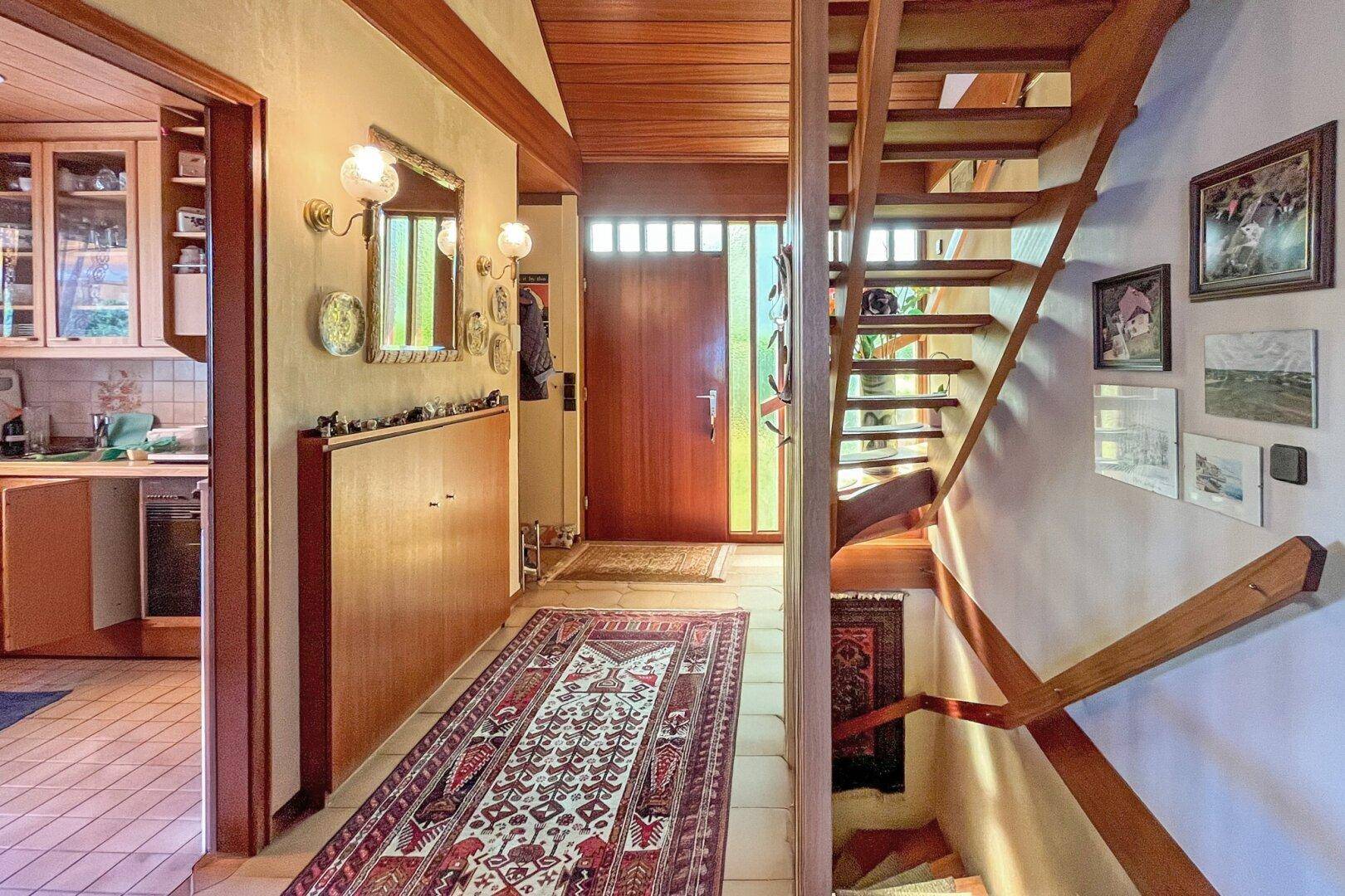 Heller Eingangsbereich mit Holztreppe, Fliesenboden und Zugang zur Küche.