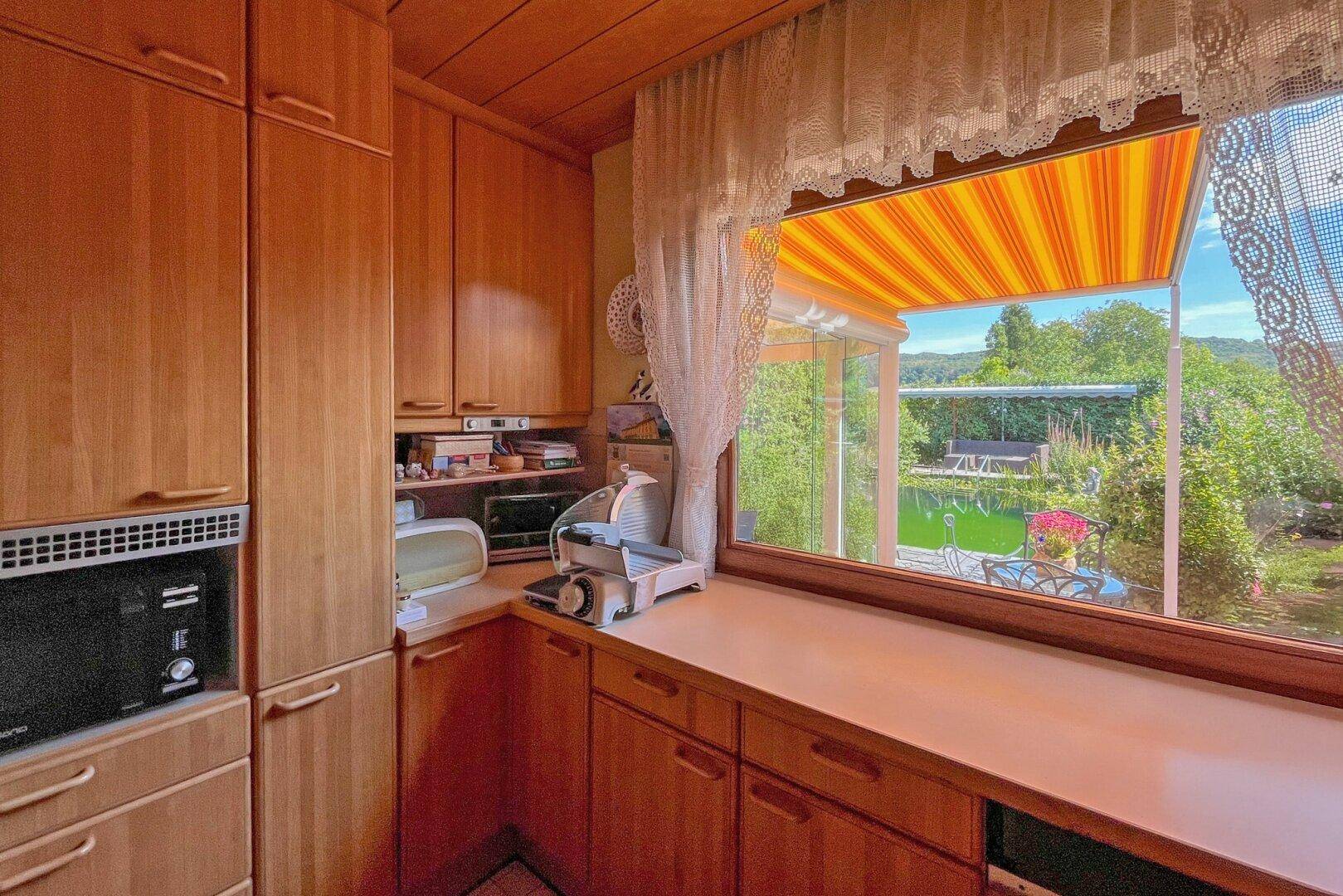 Funktionale Küche mit Holzschränken, Arbeitsplatte und Fenster mit Blick ins Grüne.