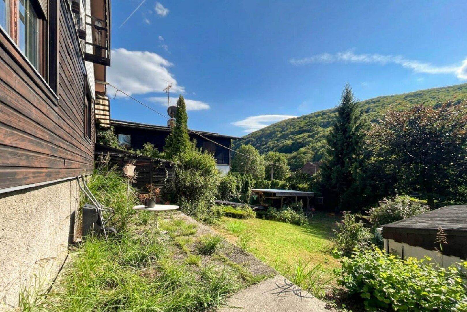 Seitlicher Gartenbereich mit kleiner Sitzgelegenheit und weitem Blick auf die grüne, hügelige Landschaft.