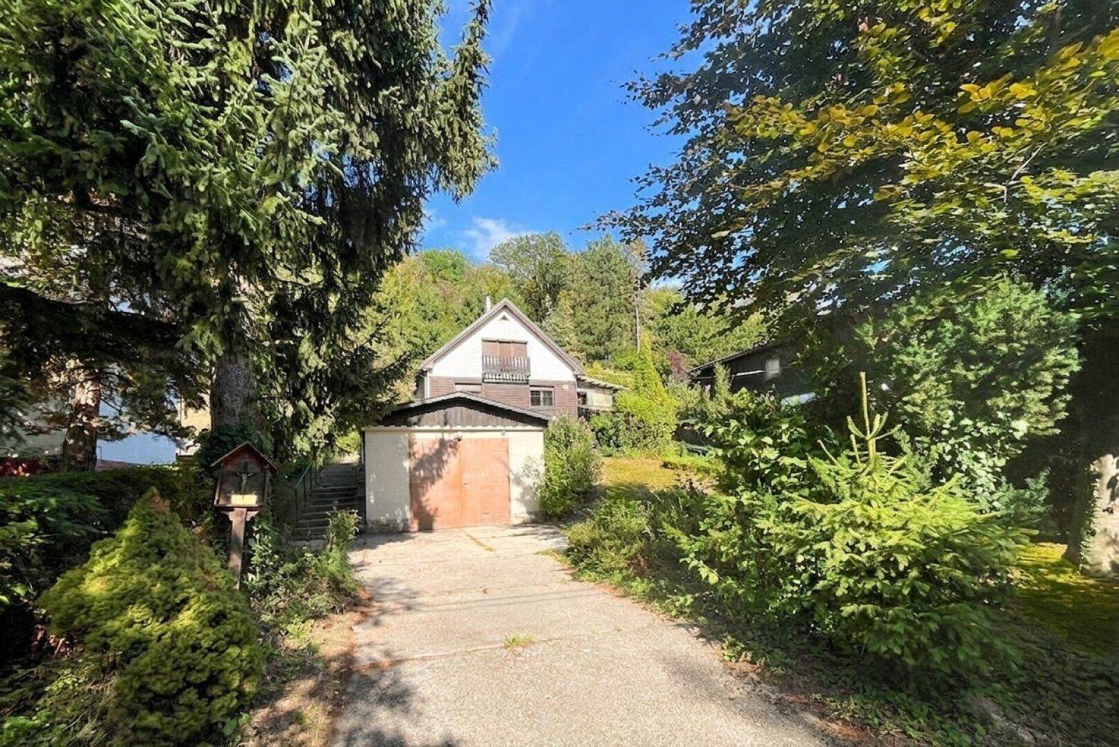 Zufahrt zum Haus mit Garage, umgeben von üppiger Vegetation und hohen Bäumen.
