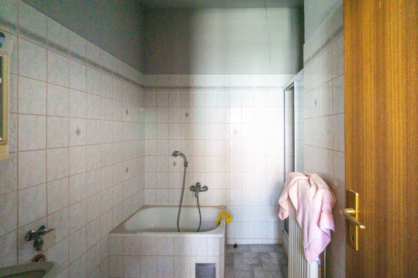 Gefliestes Badezimmer mit Badewanne und Duschvorrichtung, in renovierungsbedürftigem Zustand.