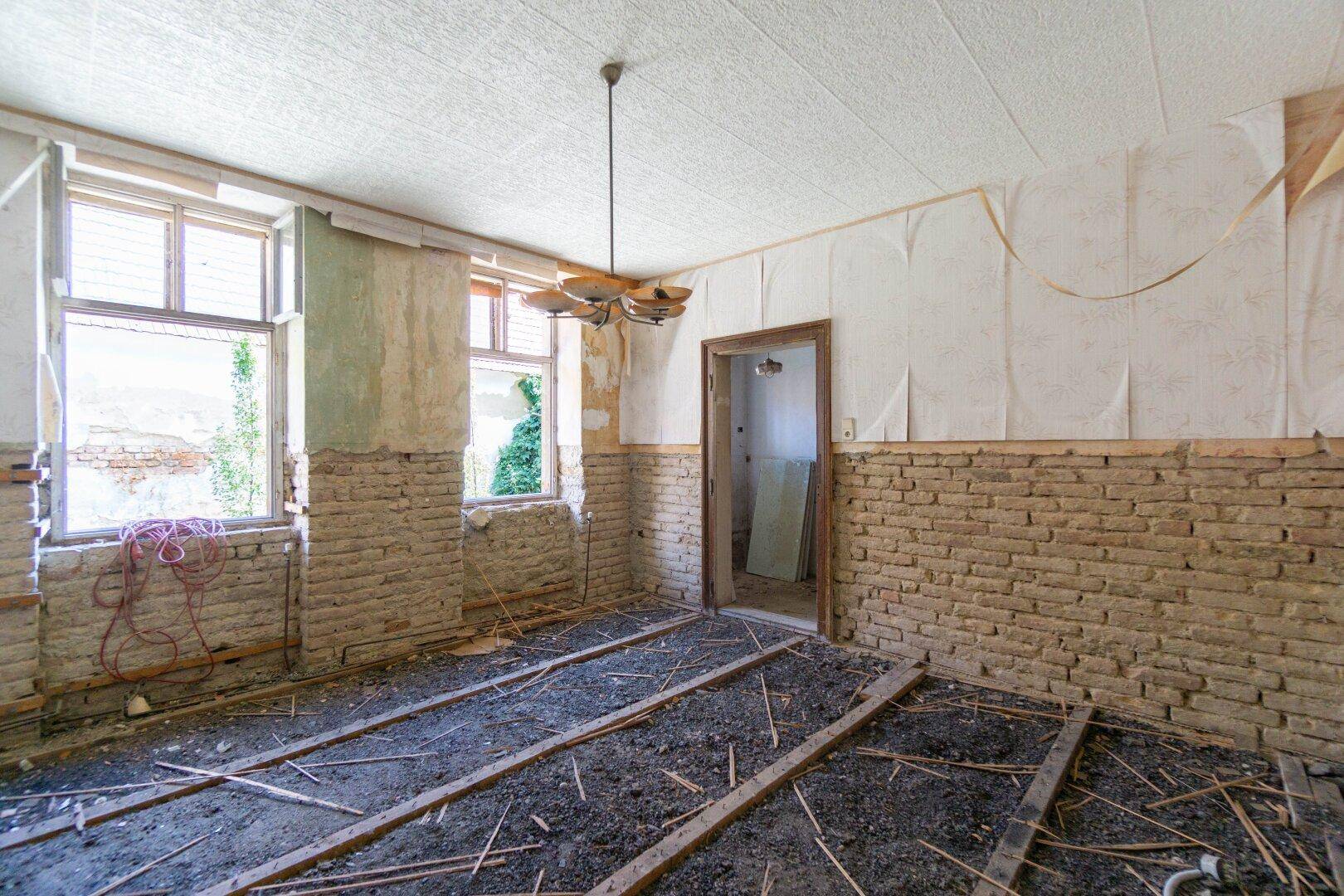 Raum in Renovierung mit freigelegten Ziegelwänden und Bodenbalken, viel natürliches Licht.