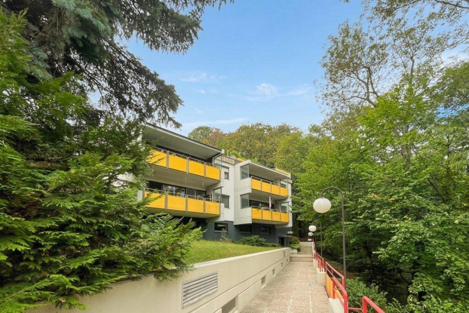 Mehrfamilienhaus mit gelben Balkonen, umgeben von viel Grün und Bäumen.