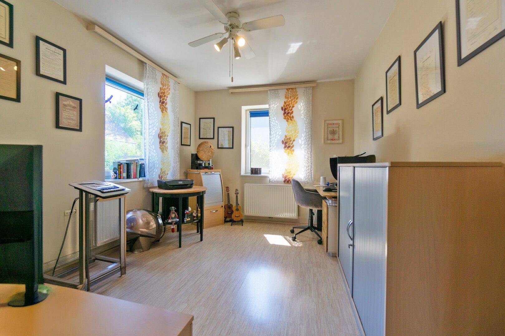 Geräumiges Arbeitszimmer mit Laminatboden, Deckenventilator und zwei Fenstern.