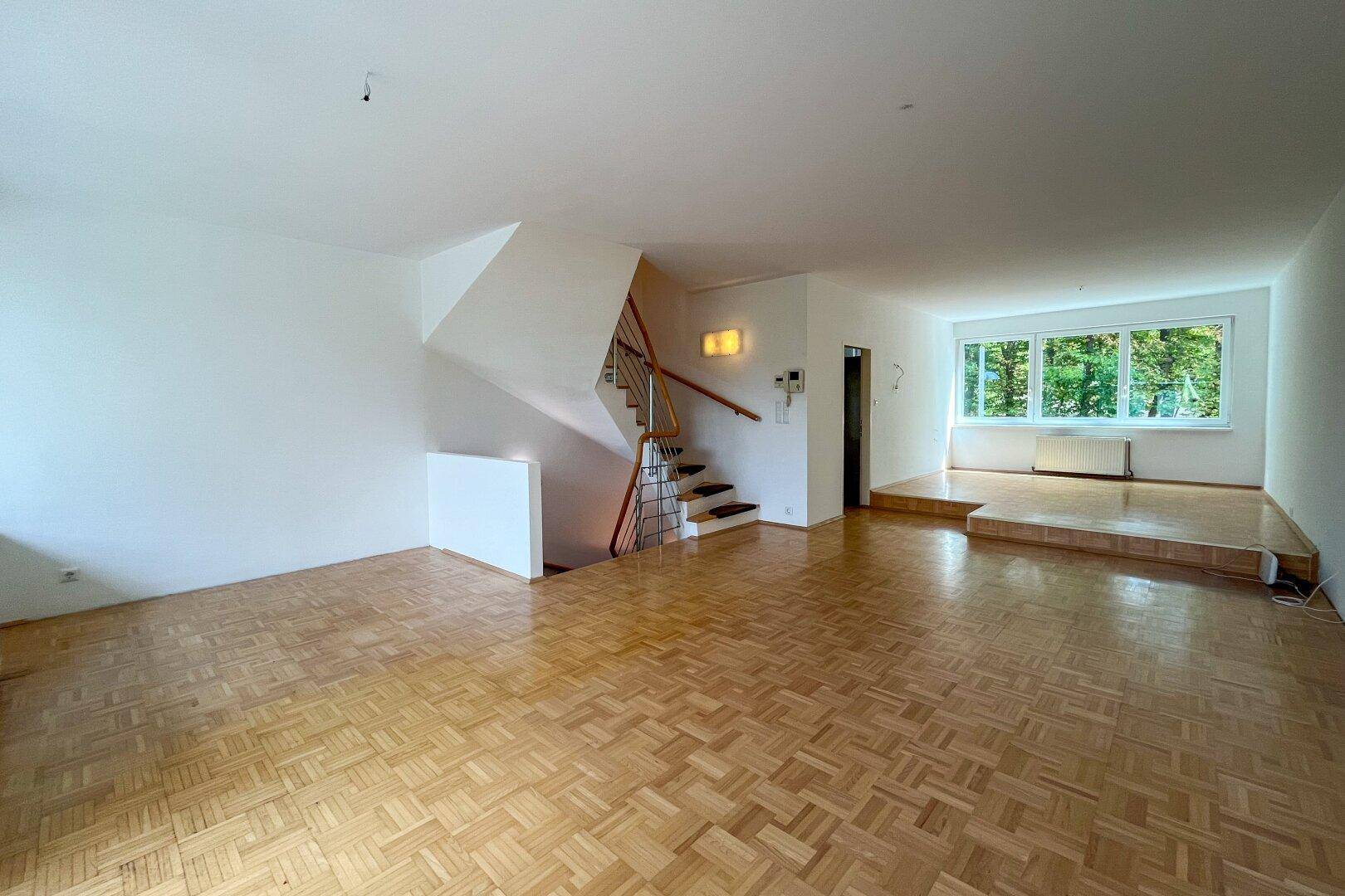 Geräumiger Wohnbereich mit Parkettboden, Treppe und erhöhter Fensterfront mit Blick ins Grüne.
