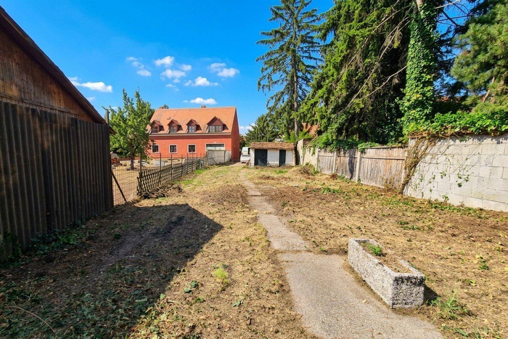 Weitläufiges, ungenutztes Grundstück mit einem Pfad und alten Nebengebäuden im Hintergrund.