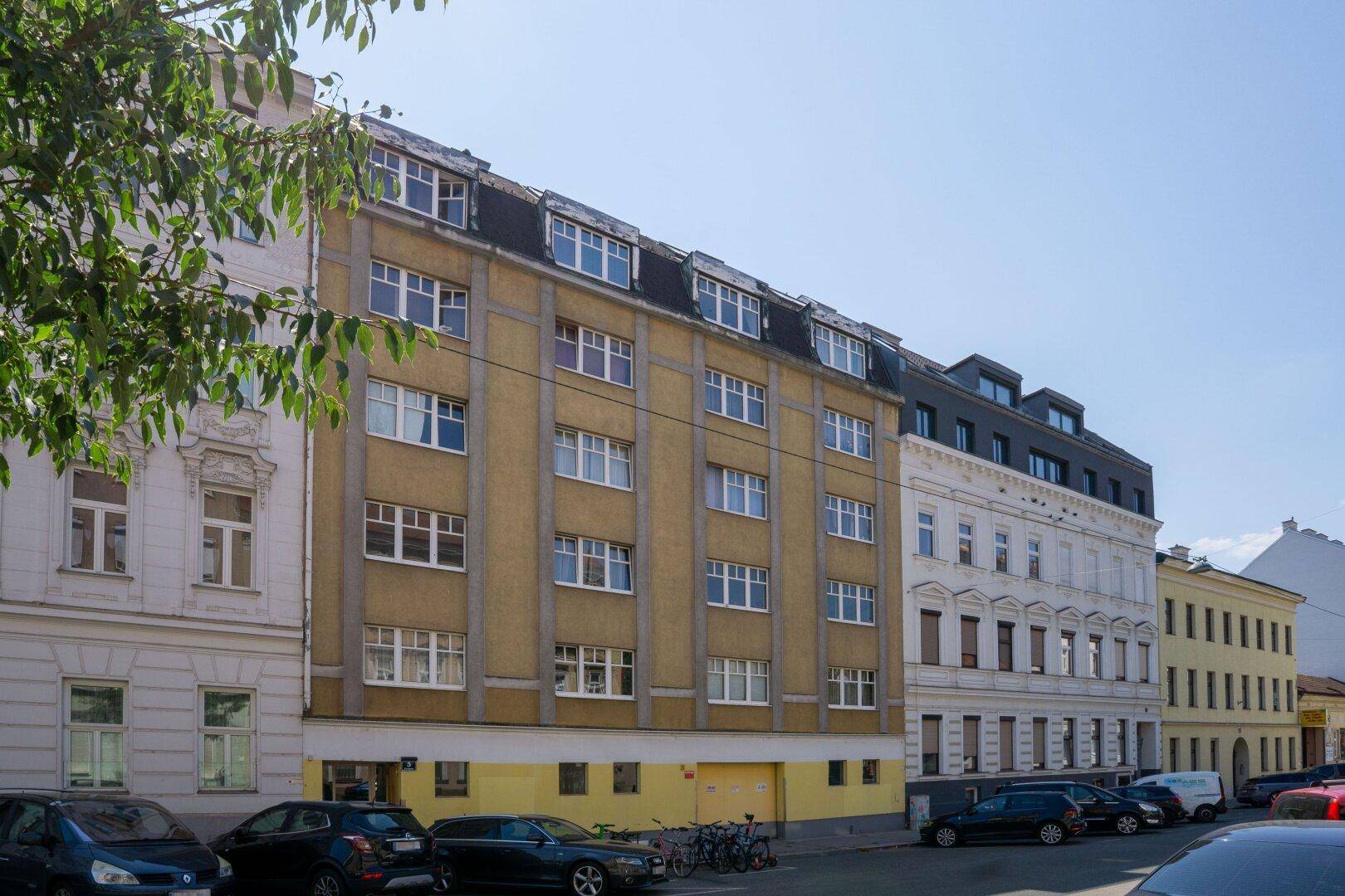 Mehrstöckiges Wohngebäude mit gelber Fassade, vielen Fenstern und parkenden Autos.