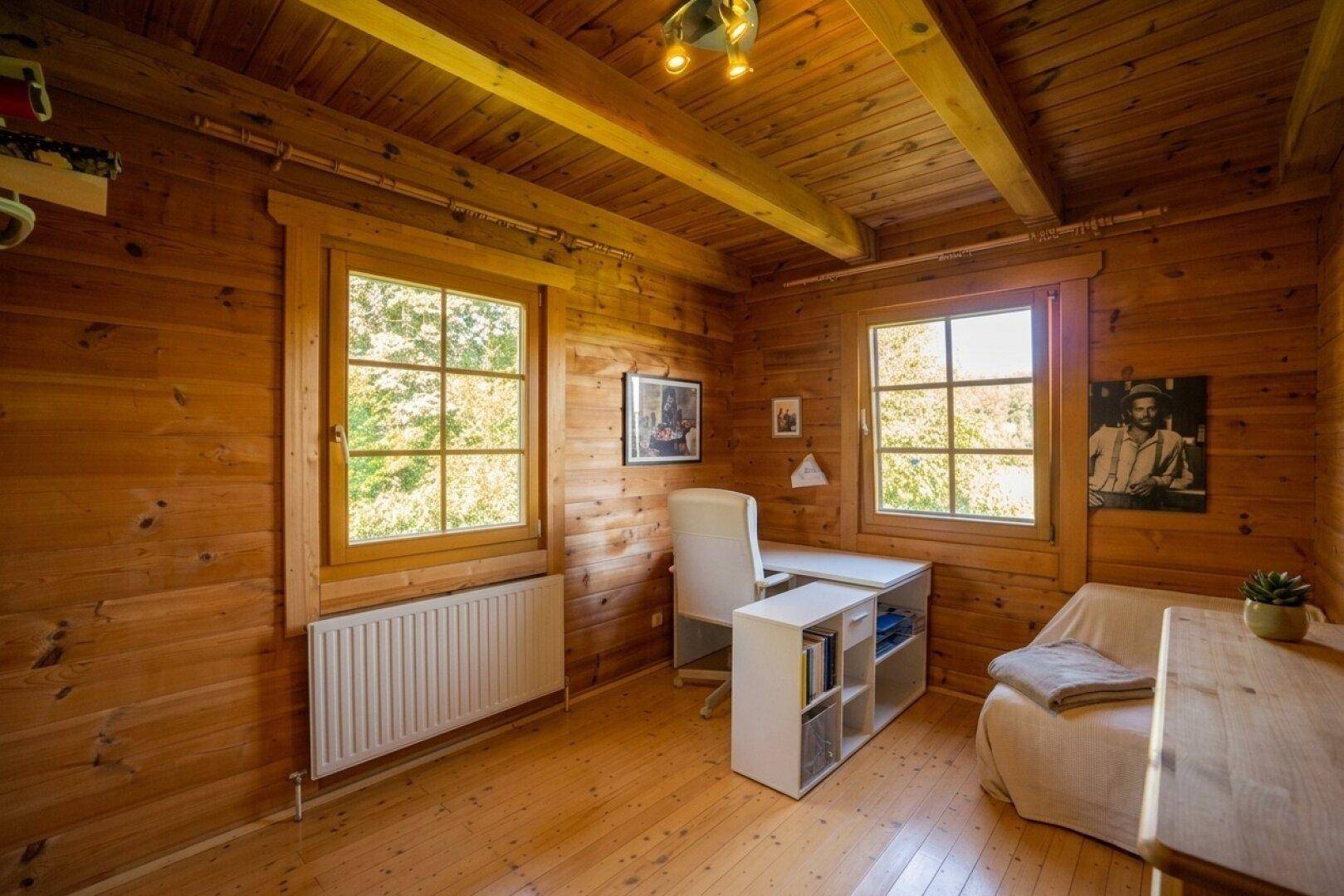 Helles Arbeitszimmer mit Holzverkleidung, Holzboden und zwei Fenstern mit Blick ins Grüne.