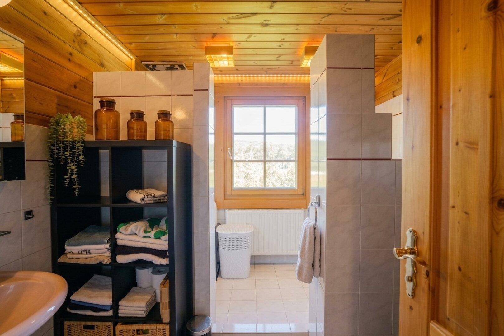 Badezimmer mit Fliesenboden, Holzdecke und Fenster für natürliche Belüftung und viel Licht.