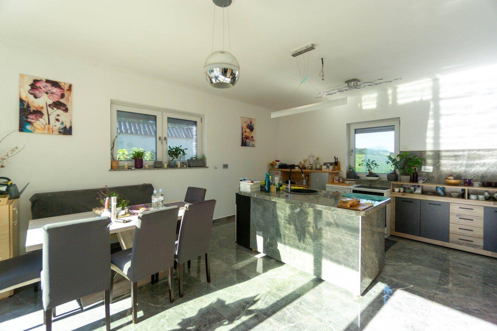 Moderne Küche mit Kücheninsel, Essbereich und viel Tageslicht durch große Fenster.