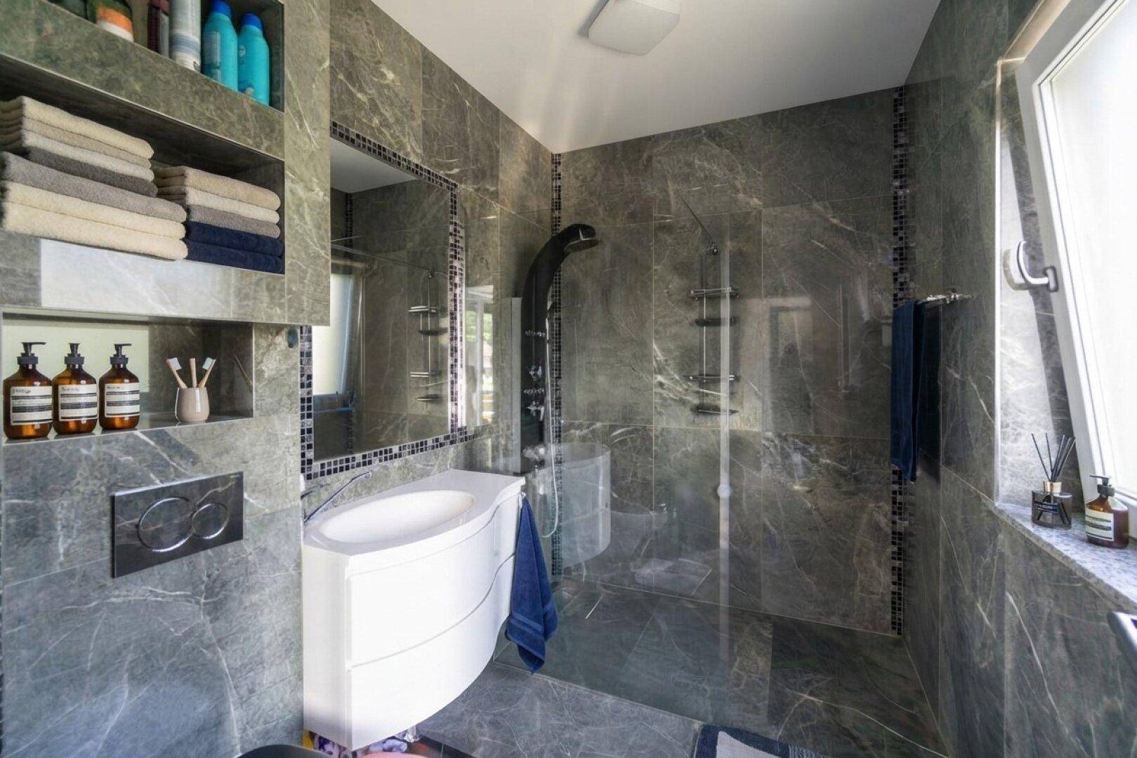 Modernes Badezimmer mit grauen Marmorfliesen, begehbarer Dusche und integrierten Ablagen.