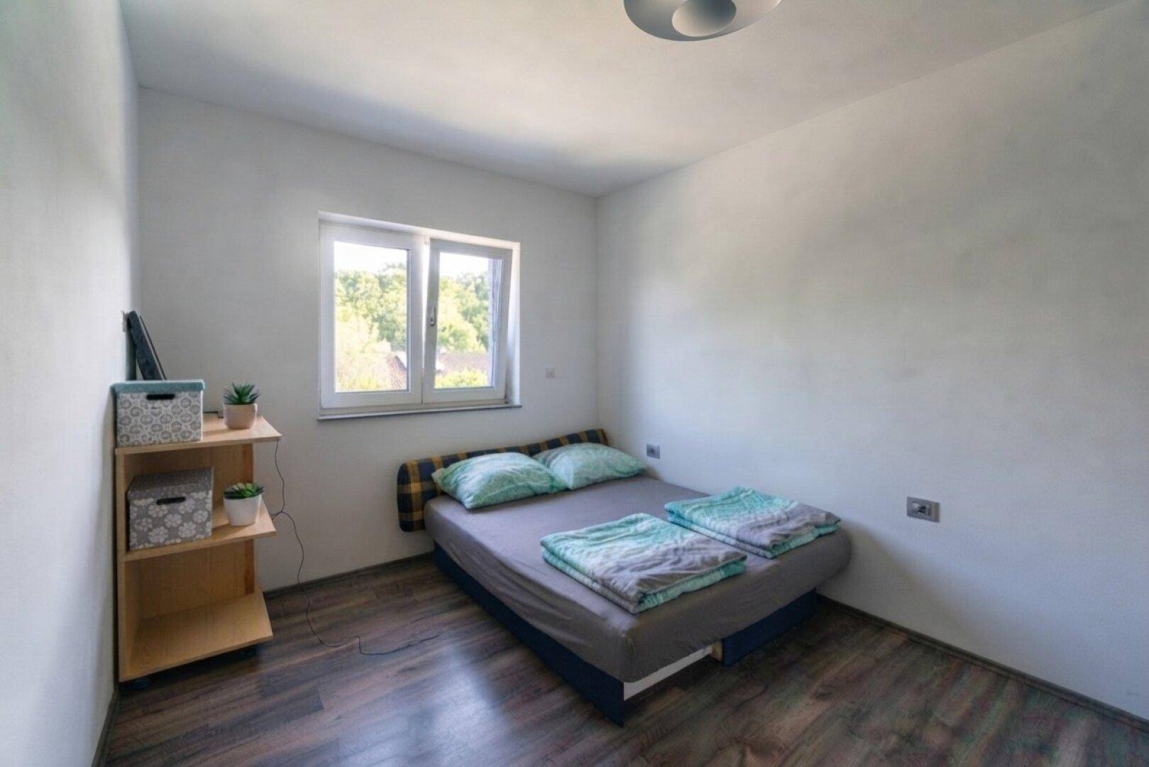 Einfaches Schlafzimmer mit Holzboden, Doppelbett und Fenster für natürliches Licht.