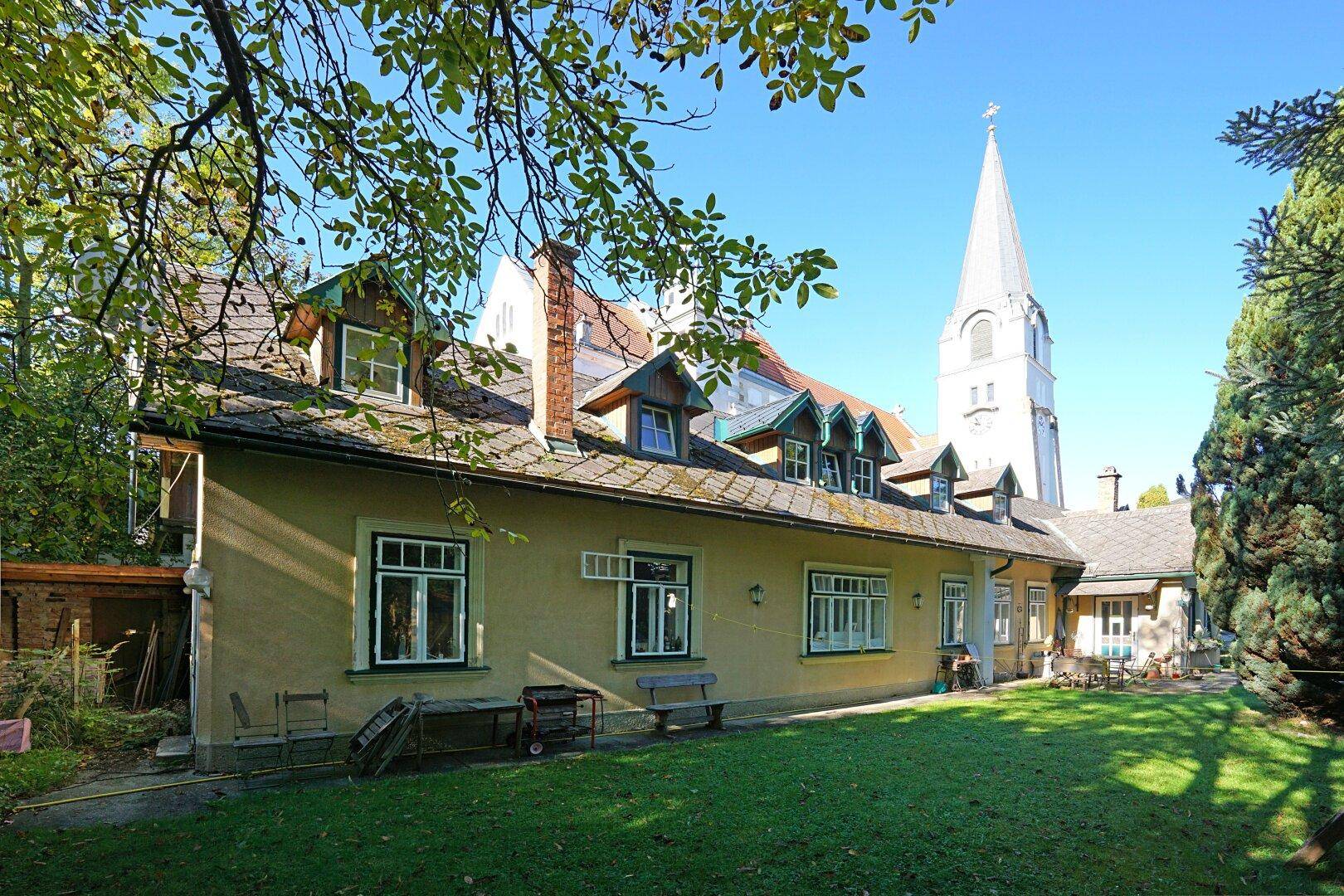 Seitenansicht des charmanten Hauses mit grünem Garten und dem Kirchturm im Hintergrund.