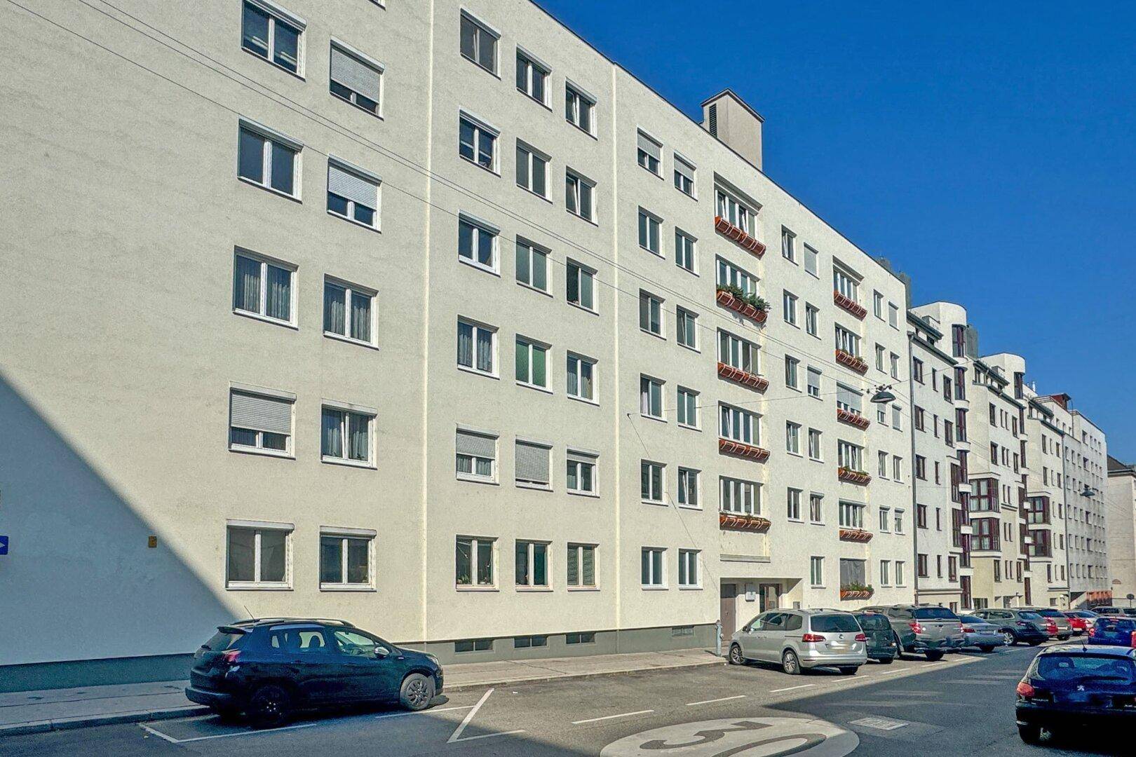 Mehrfamilienhaus mit heller Fassade und vielen Fenstern an einer städtischen Straße.
