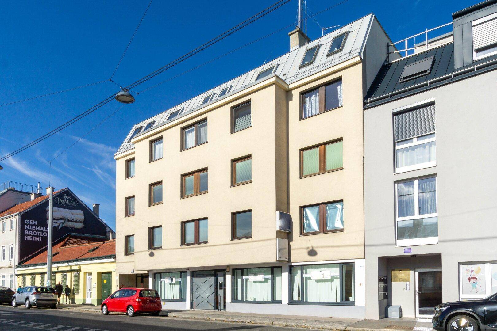Mehrstöckiges Wohngebäude mit beiger Fassade und Mansarddach an einer städtischen Straße.