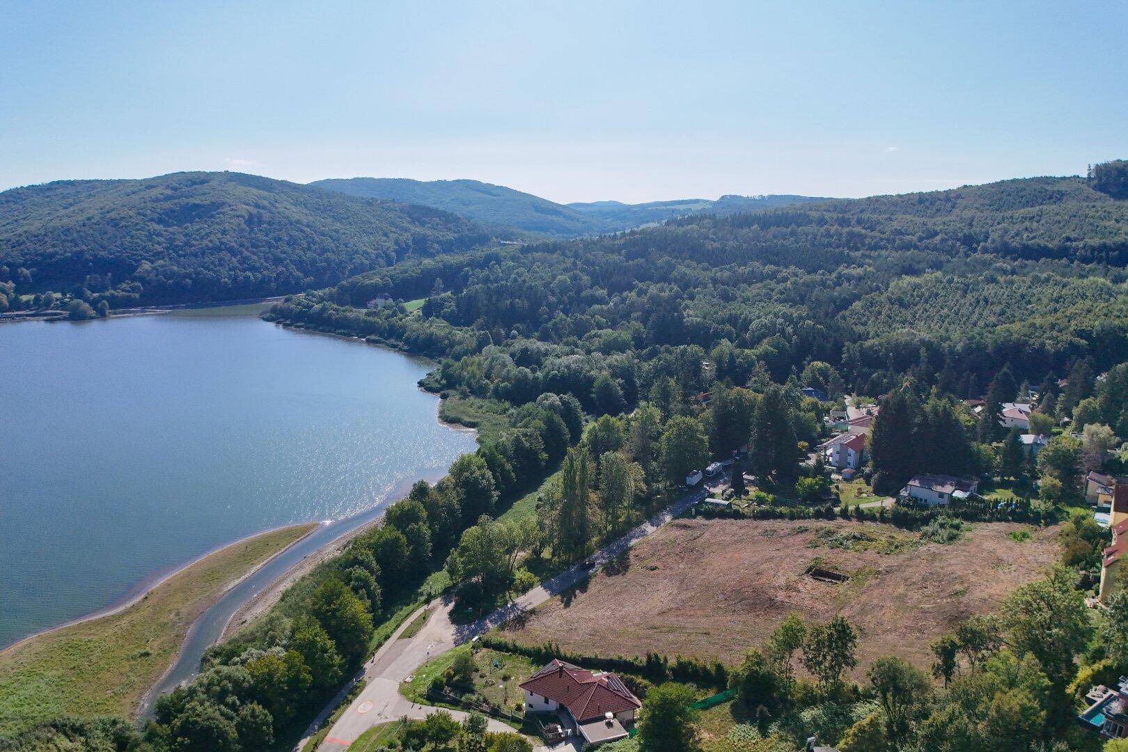 Weitläufige Luftaufnahme mit einem See, bewaldeten Hügeln und Häusern am Ufer.