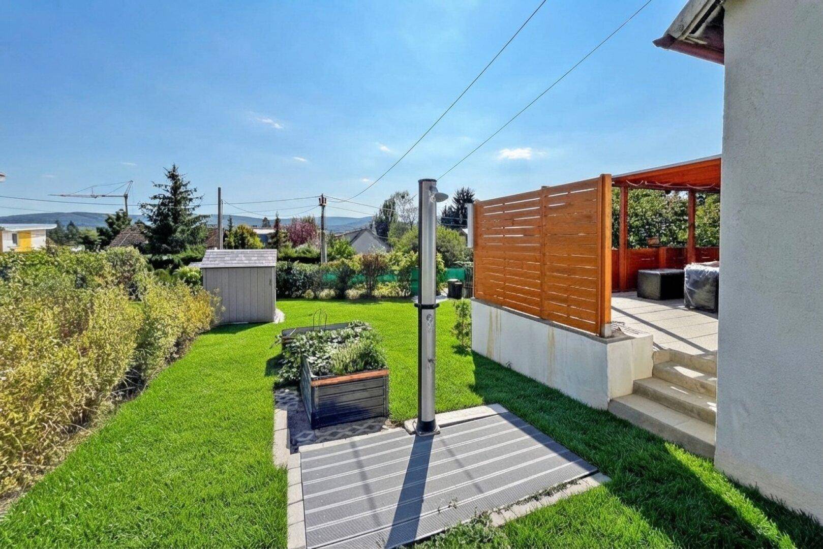 Grüner Garten mit Außendusche und Blick auf die angrenzende Holzterrasse.