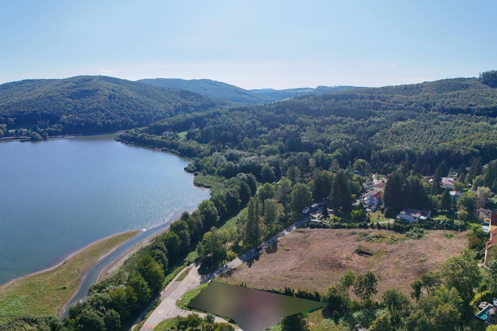 Luftaufnahme eines großen Grundstücks am Seeufer mit bewaldeten Hügeln im Hintergrund.