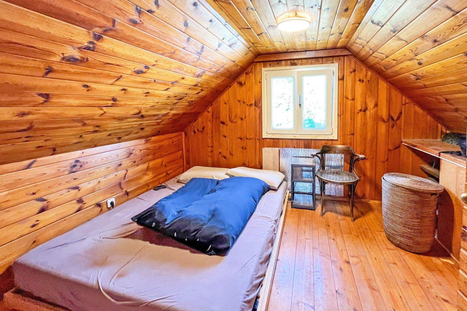 Schlafzimmer im Dachgeschoss mit Holzwänden, Bett und Fenster unter der Dachschräge.