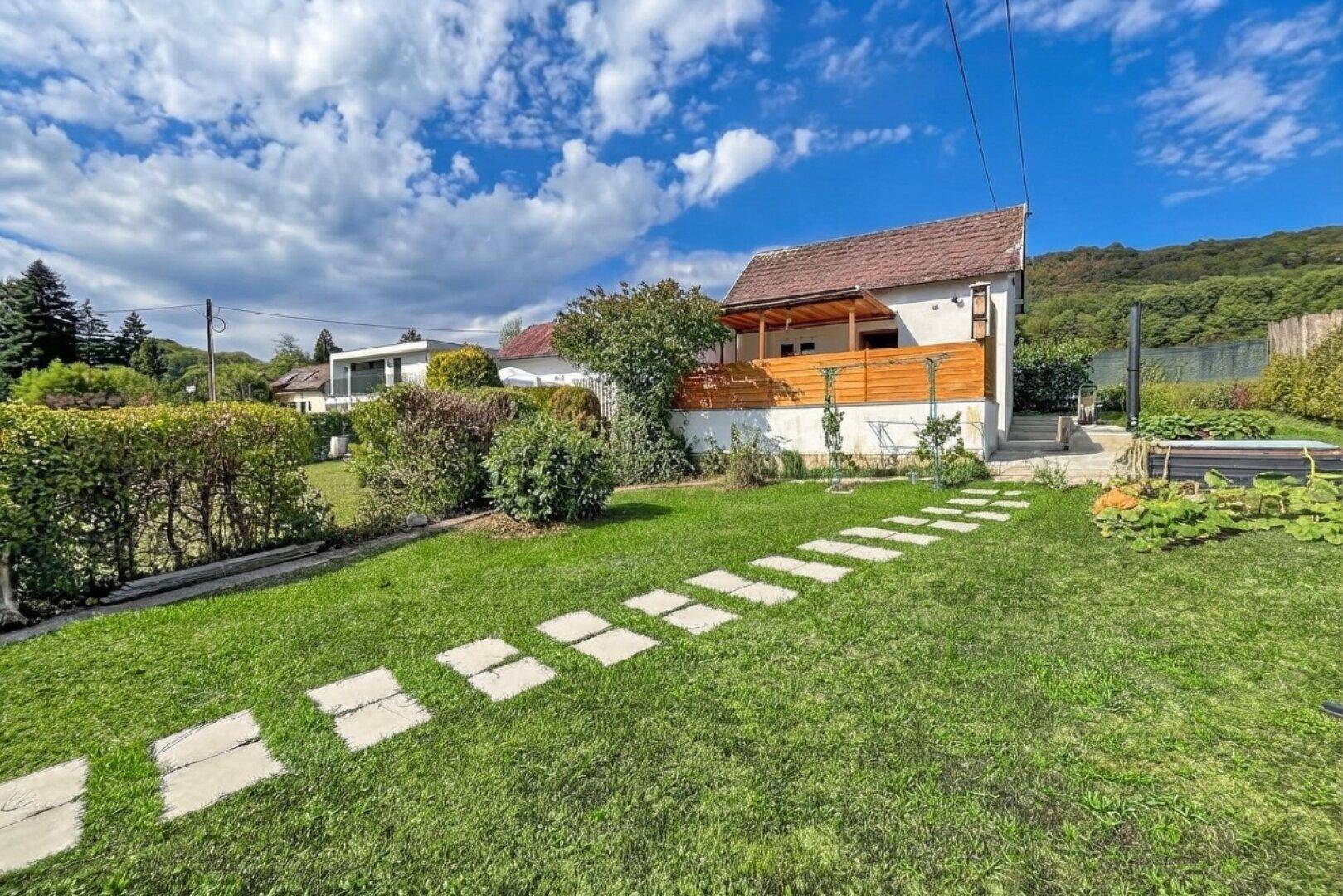 Hausfassade mit Holzterrasse und gepflegtem Garten mit Steinweg und Rasenfläche.