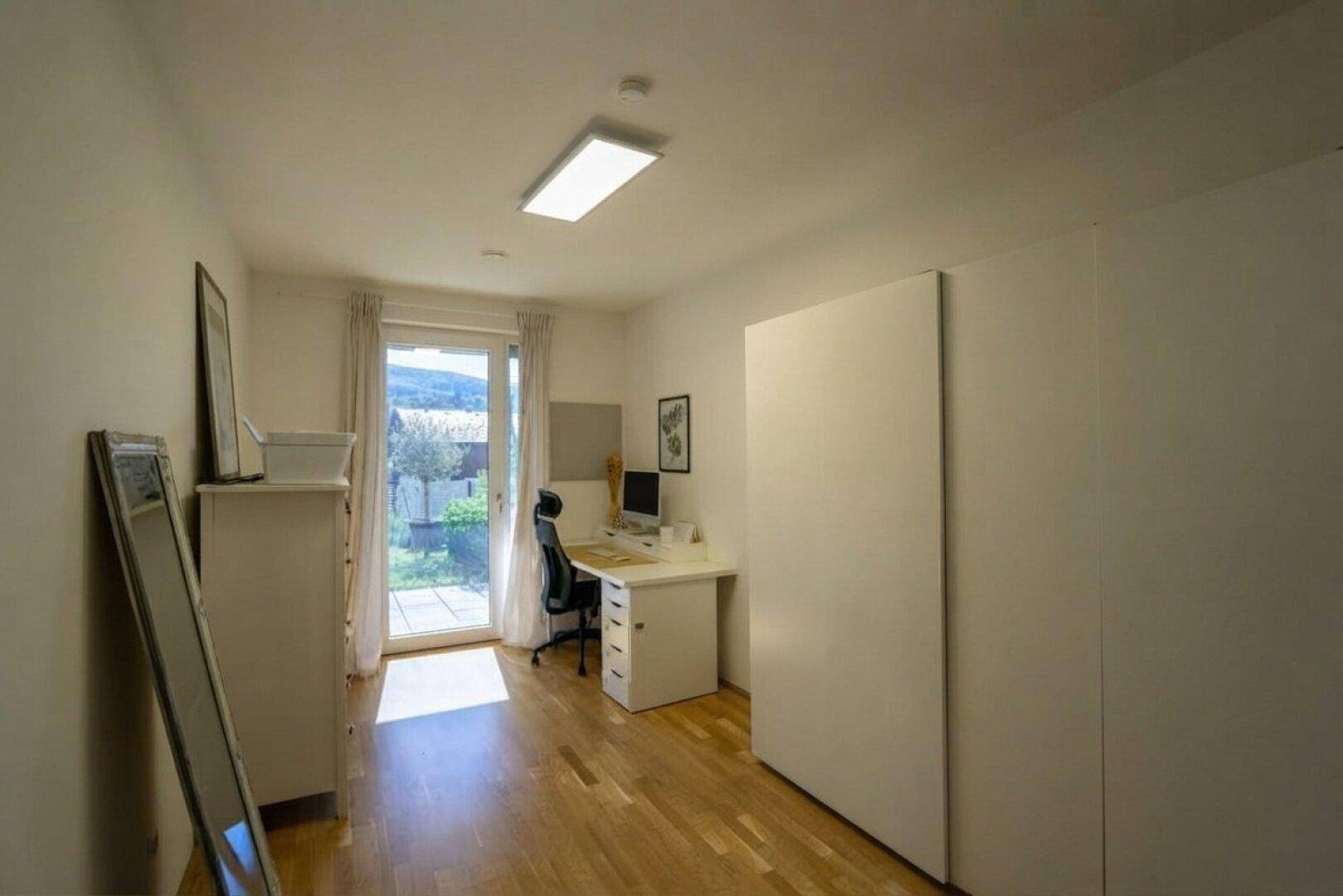 Helles Arbeitszimmer mit Holzboden, Schreibtisch und Zugang zu einem Außenbereich.