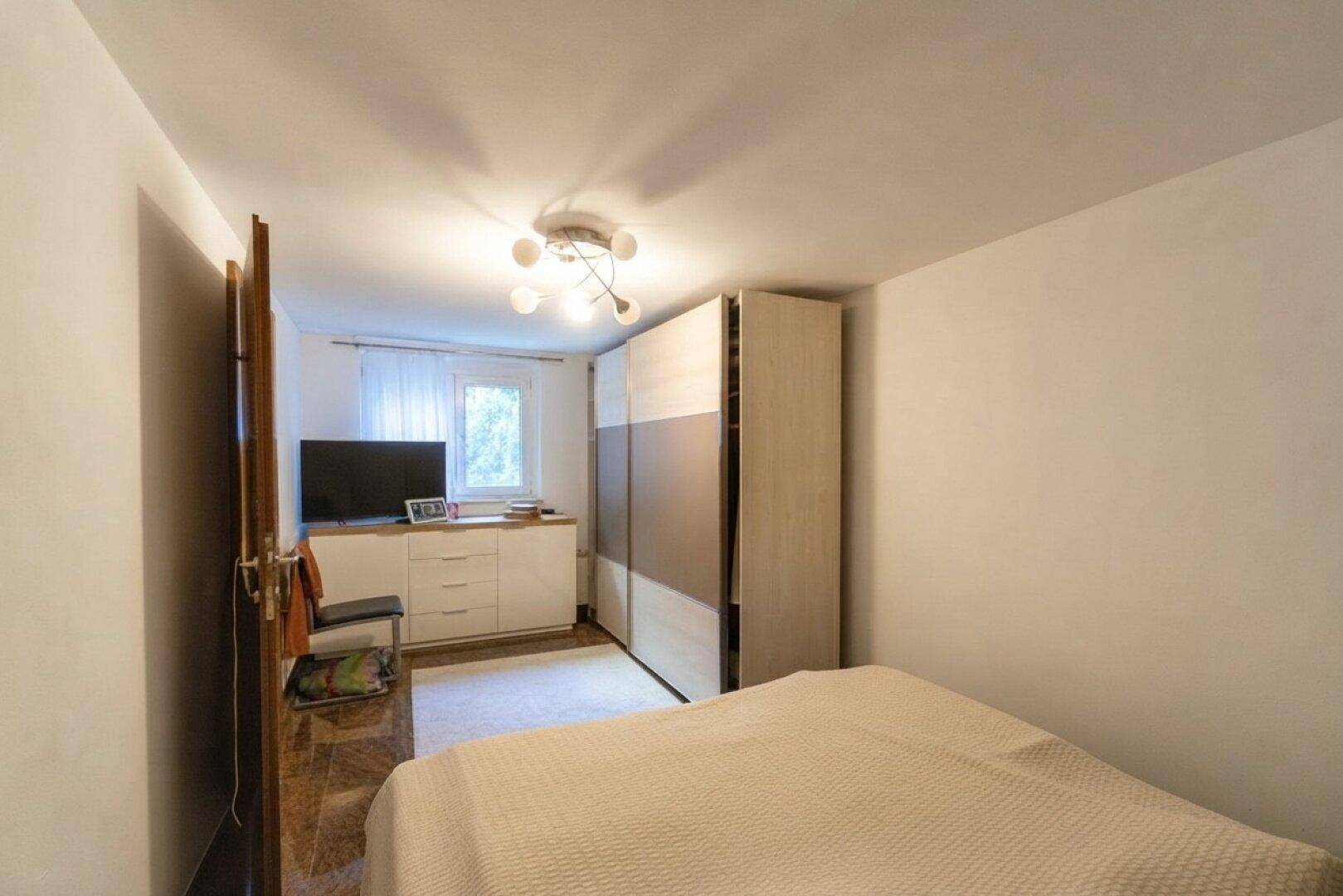 Schlafzimmer mit Bett, Kleiderschrank und Fenster, das viel Tageslicht hereinlässt.
