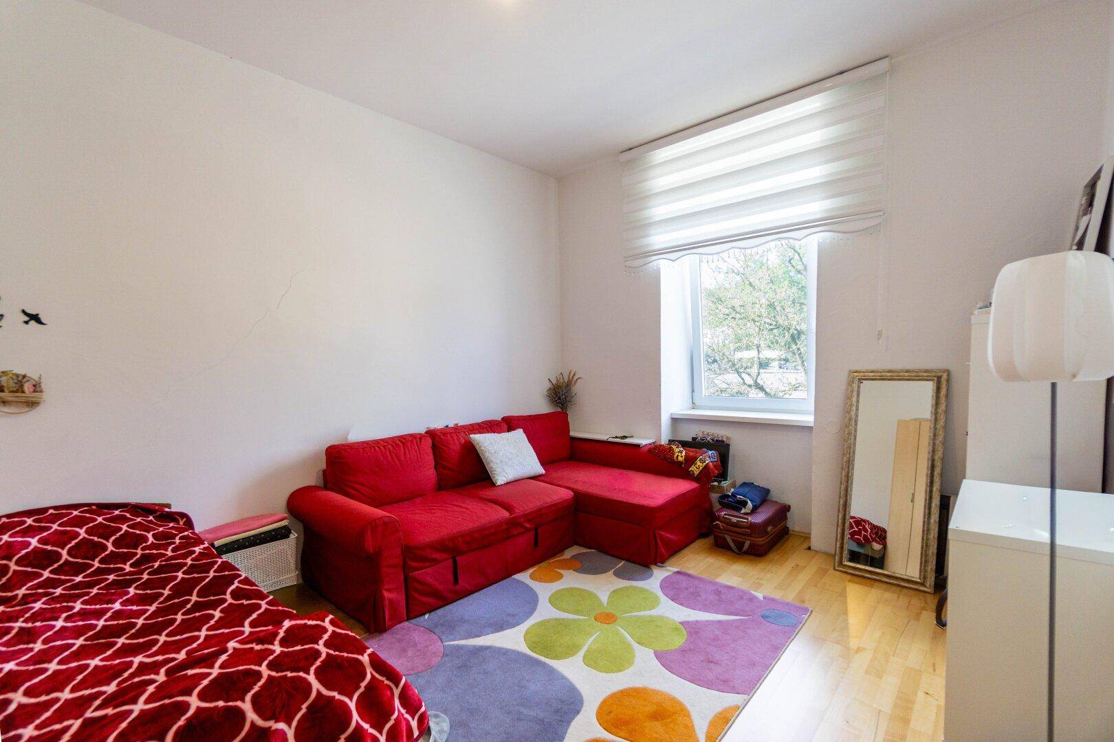 Gemütliches Schlafzimmer mit rotem Sofa, Holzboden und einem großen Fenster für viel Tageslicht.