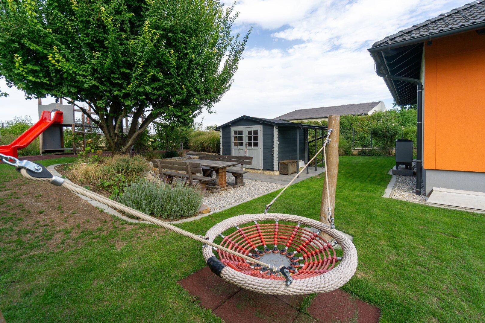 Großer Garten mit Spielplatz, Schaukel, Rutsche und einem Gartenhaus unter einem Baum.