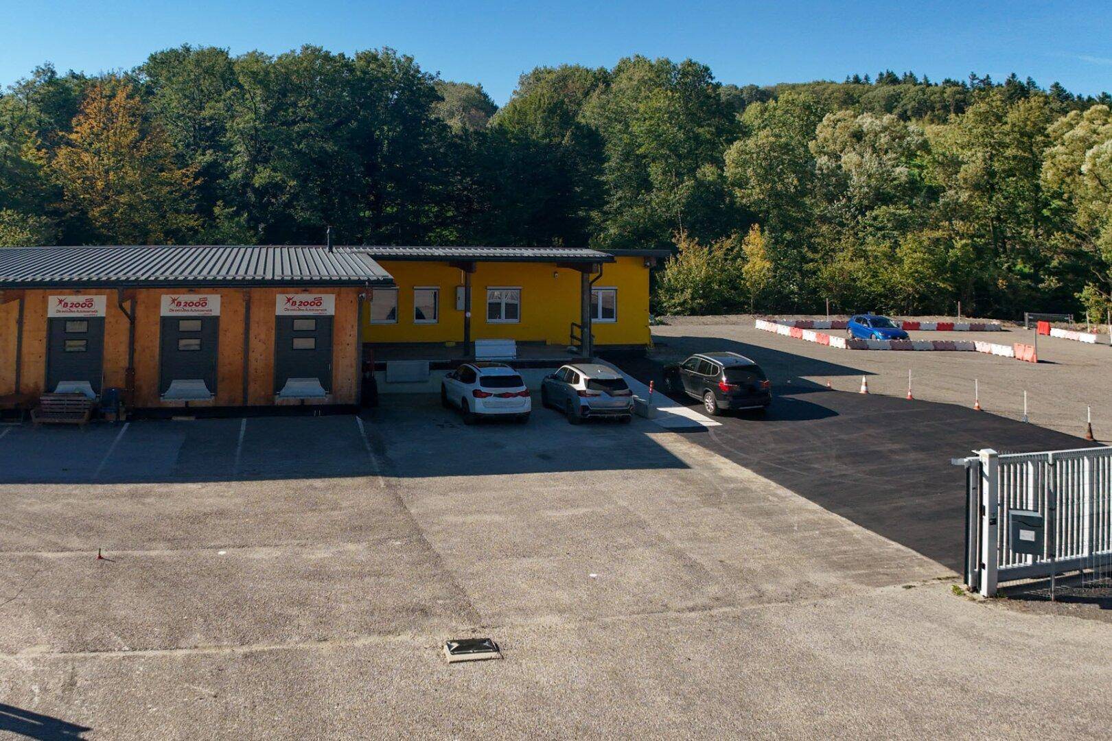 Ansicht des Gewerbegebäudes mit Holzverkleidung und gelber Fassade, umgeben von Parkplätzen und Bäumen.