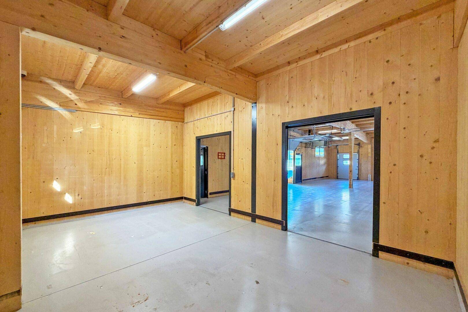 Innenraum der Lagerhalle mit Holzwänden und -decke, Betonboden und Zugang zu weiteren Räumen.
